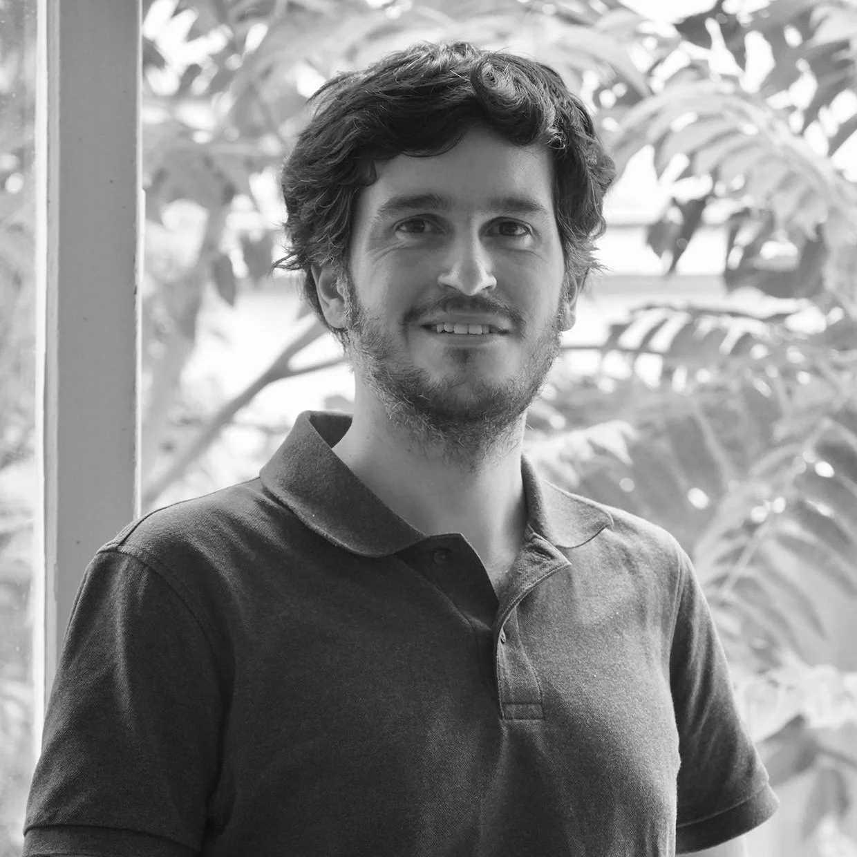 Portrait d'un homme avec des cheveux bouclés, une barbe et un sourire, portant un polo, dans un environnement intérieur avec des plantes en arrière-plan.