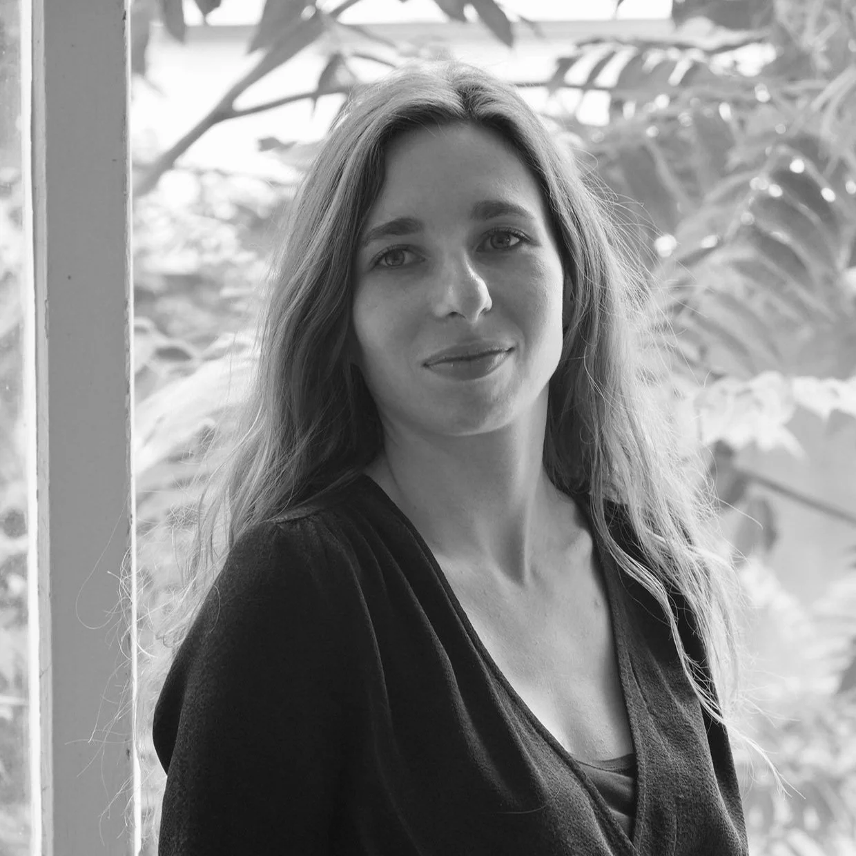 Photo en noir et blanc d'une femme aux cheveux longs et ondulés, portant un haut sombre, dans un environnement naturel avec des feuilles en arrière-plan.