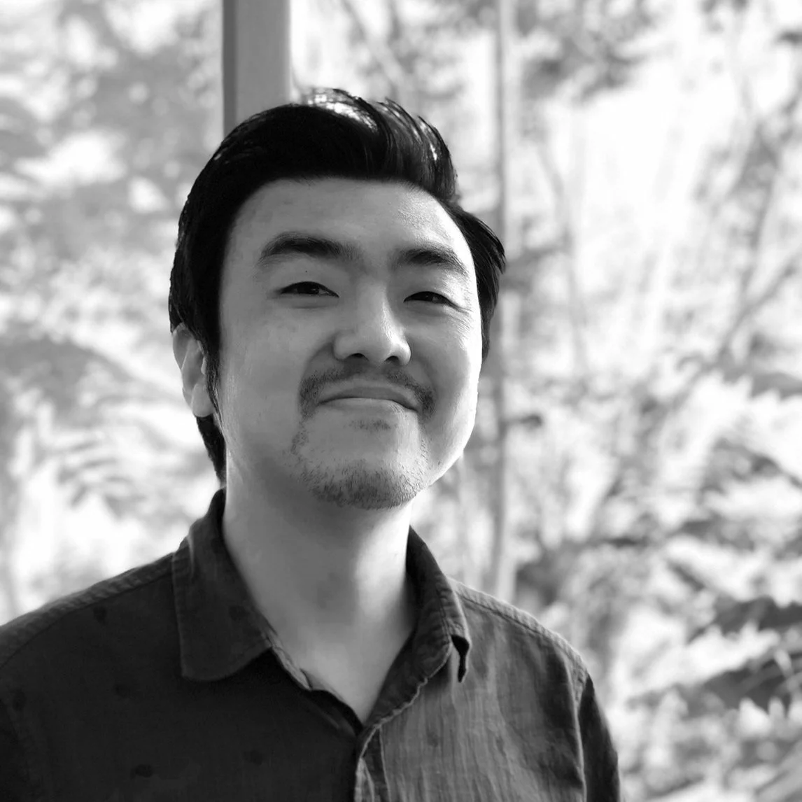 Un homme souriant en noir et blanc, portant une chemise à col, avec une fenêtre derrière lui montrant un paysage flou.