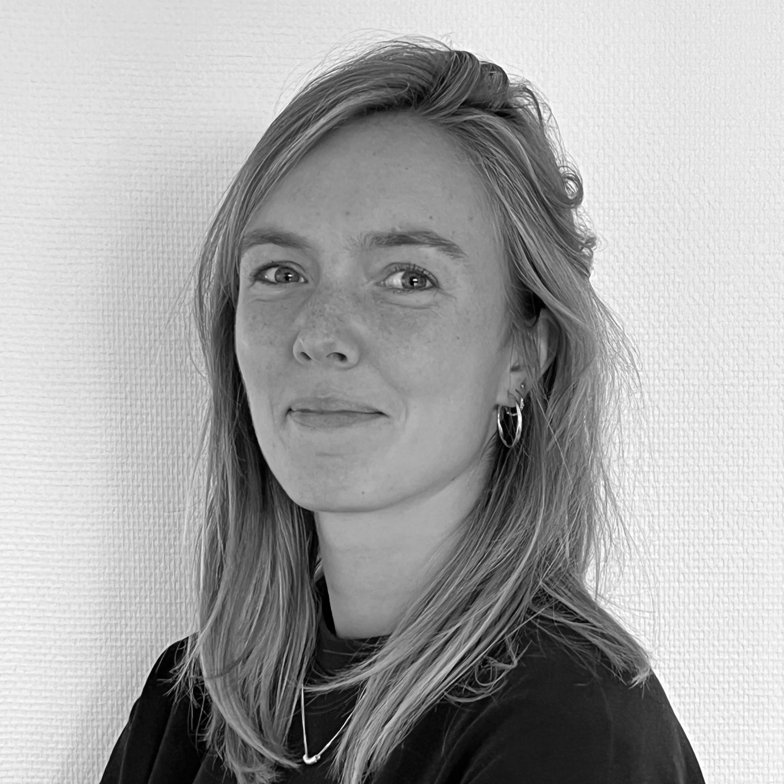 Portrait d'une femme avec des boucles d'oreilles en argent, portant un chignon léger et un haut noir, contre un fond blanc texturé.