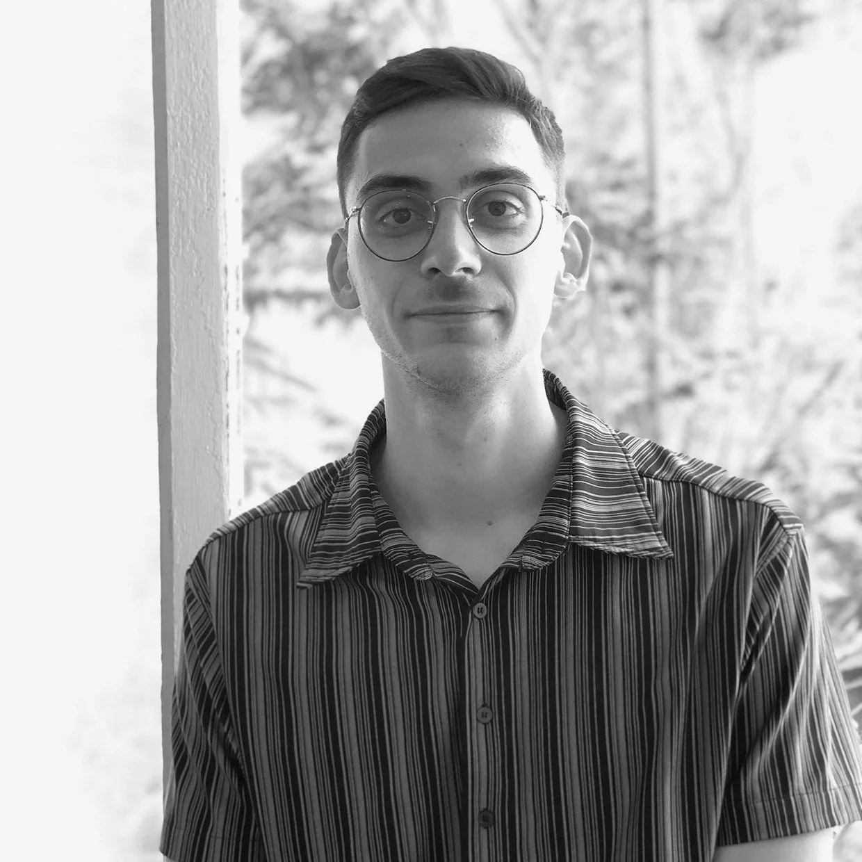 Jeune homme portant des lunettes rondes, chemise à rayures, debout près d'une fenêtre avec un arrière-plan flou de nature.