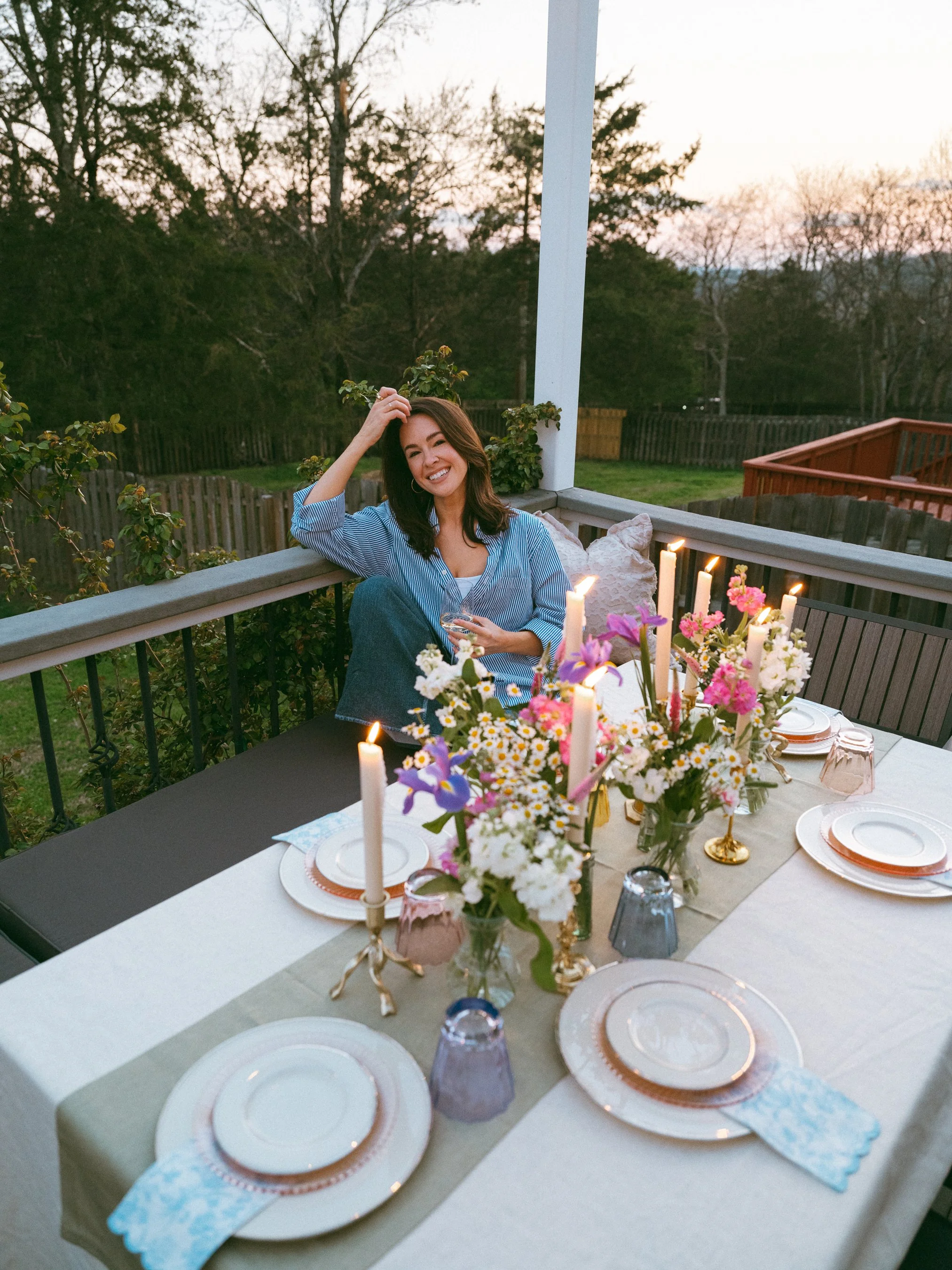 A woman sitting on a porch at sunset, smiling, with a dining table set with floral arrangements and candles in the foreground.