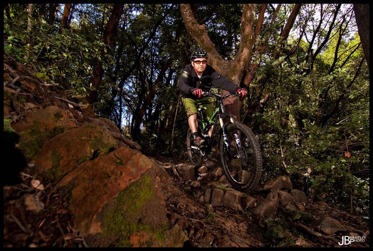 Man mountain biking on a rocky forest trail surrounded by trees and greenery.