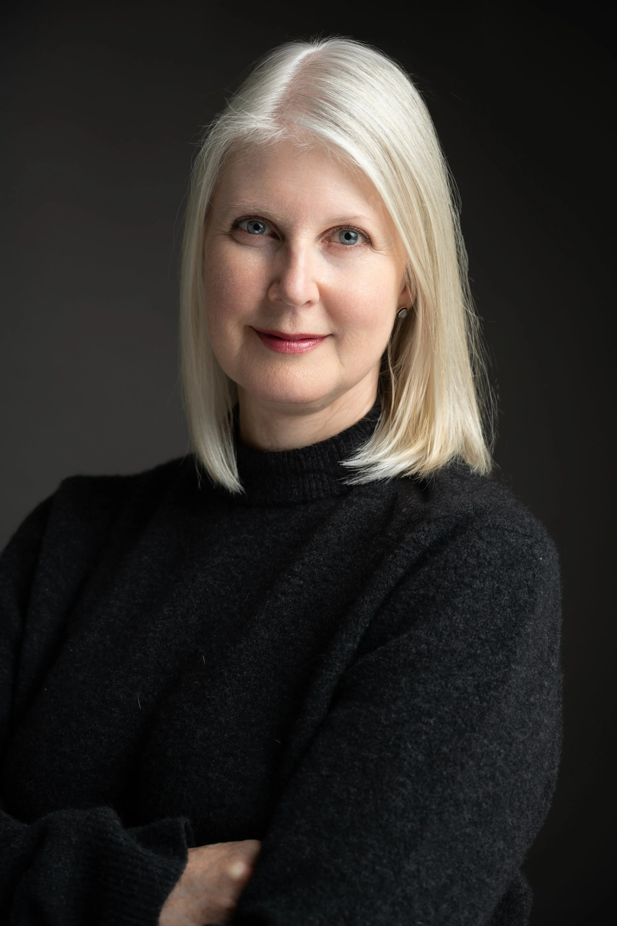 Portrait of an older woman with light skin, straight silver hair, and blue eyes, wearing a black turtleneck sweater, against a dark background.