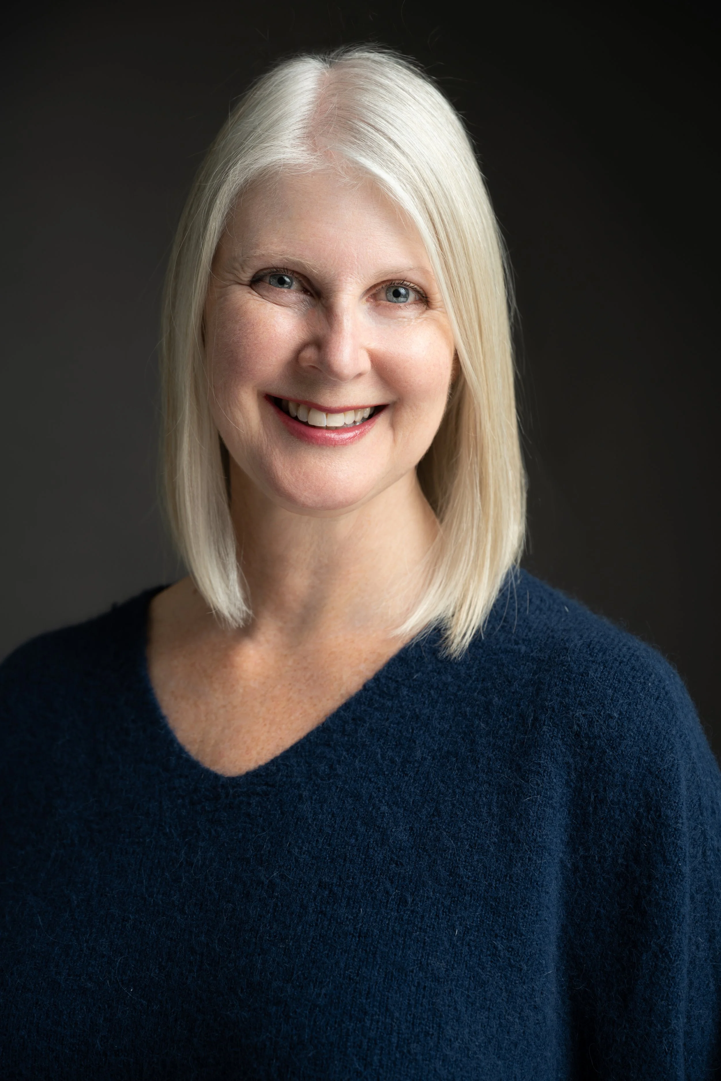 A smiling woman with blond hair, wearing a dark blue sweater, against a dark background.