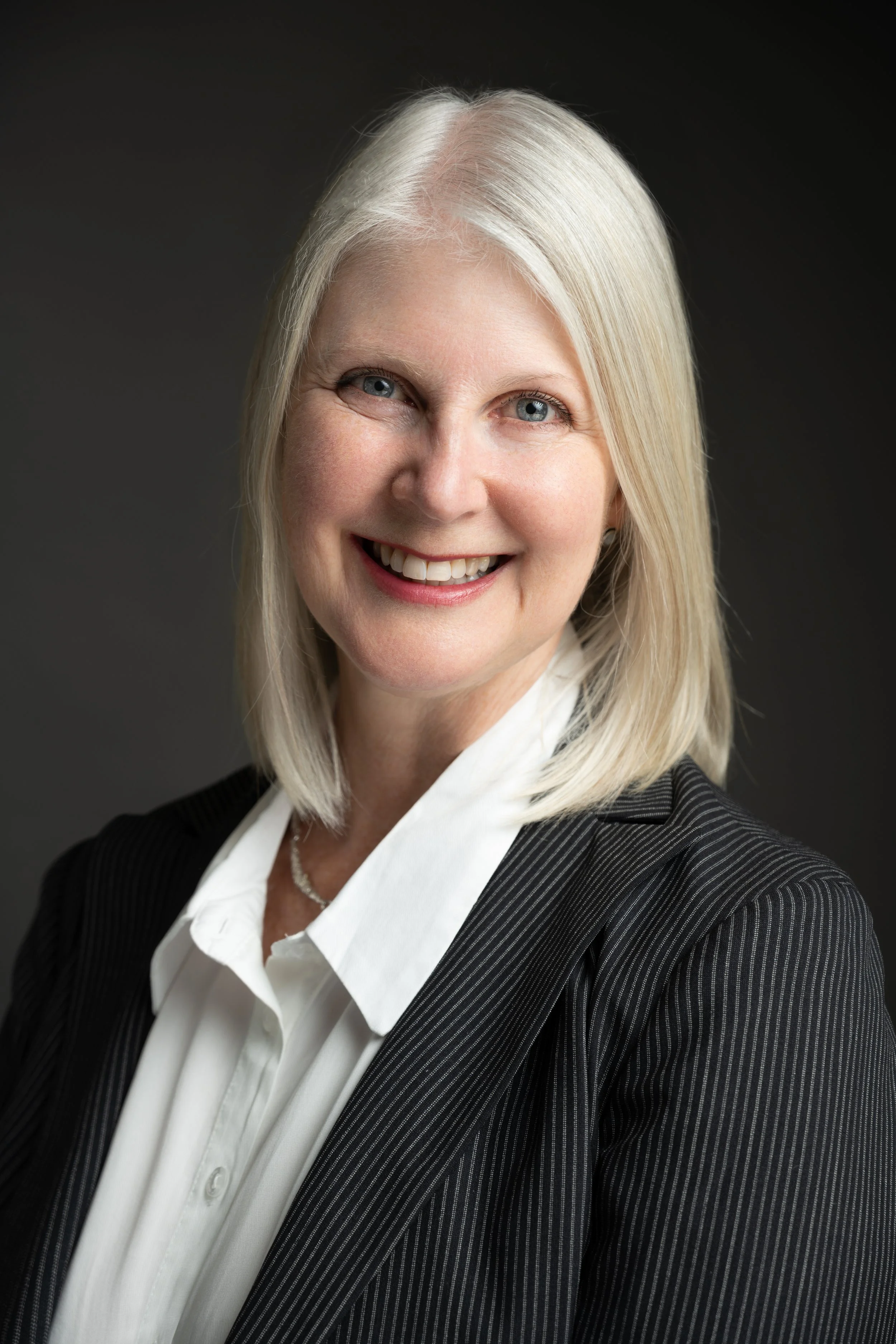 A smiling woman with shoulder-length blonde hair, wearing a black pinstripe blazer over a white blouse, against a dark background.