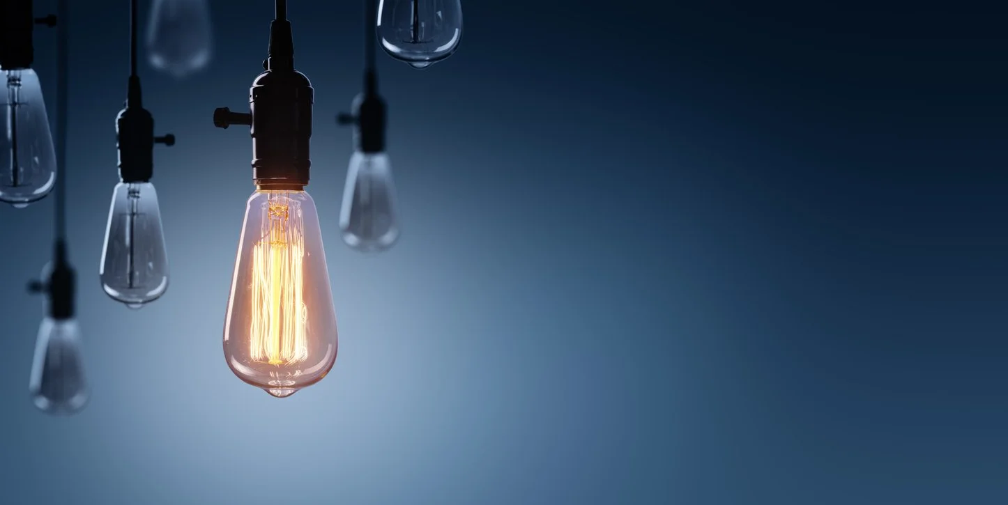 Glowing vintage Edison light bulb hanging among unlit bulbs against a dark blue background