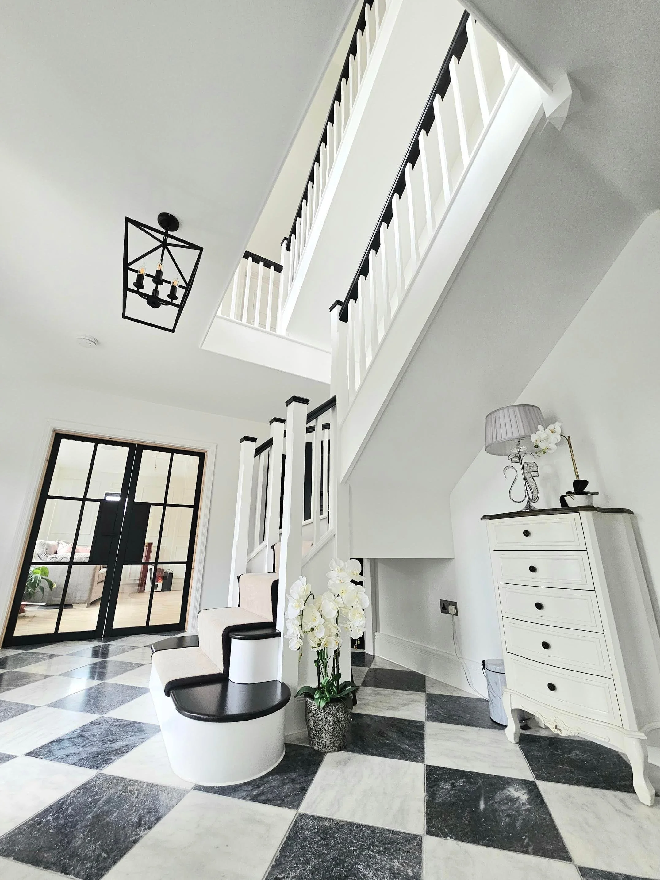 Interior of a modern home with a black and white checkered marble floor, white walls, a white staircase with black accents, a black chandelier, a white dresser with black handles, a floral table lamp, a white chair with black accents, and a potted plant with white flowers.
