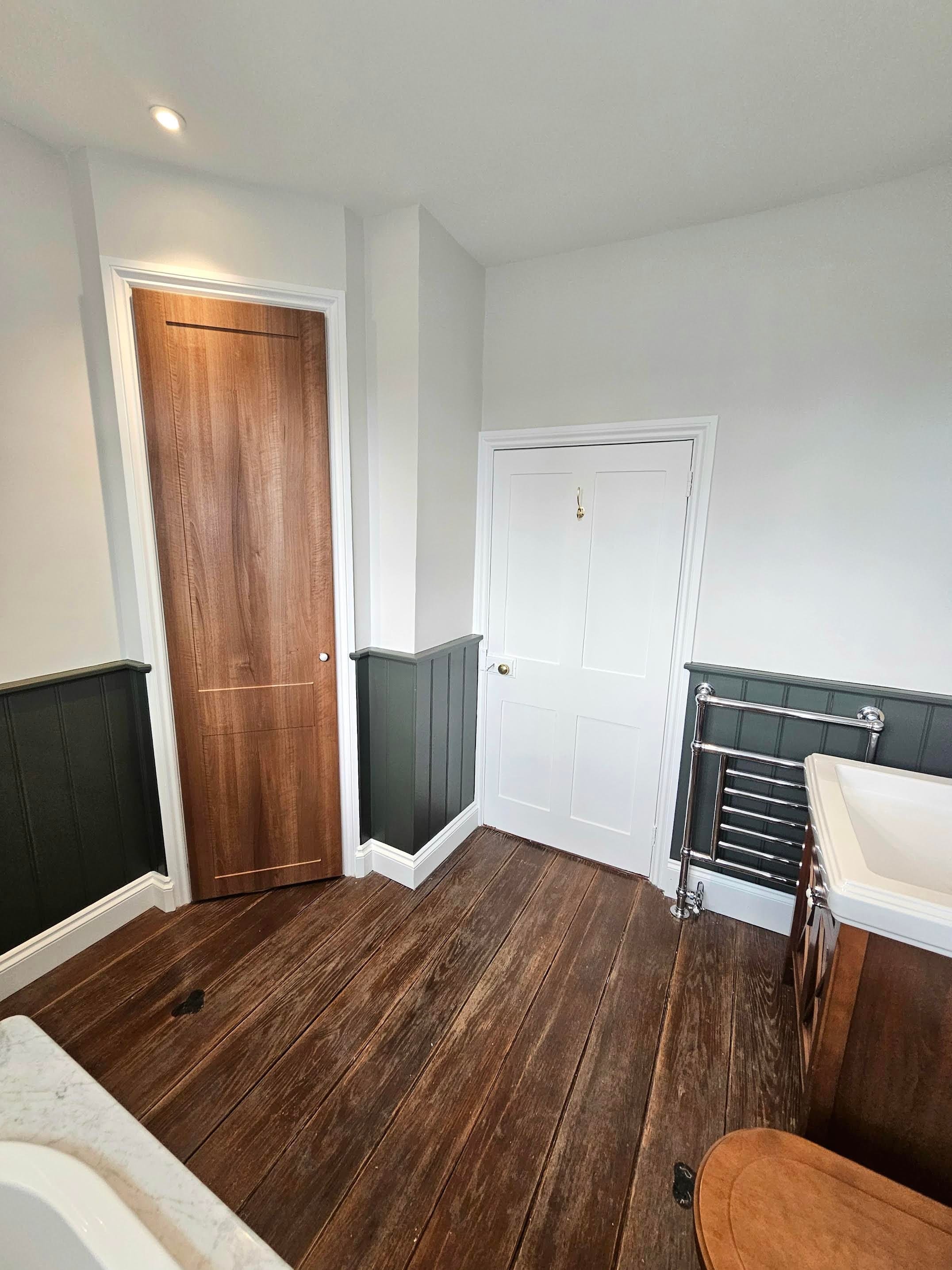 An interior bathroom scene featuring brown wooden flooring, a white door, a small closet door, a metal towel rack, and part of a white sink with a marble countertop.