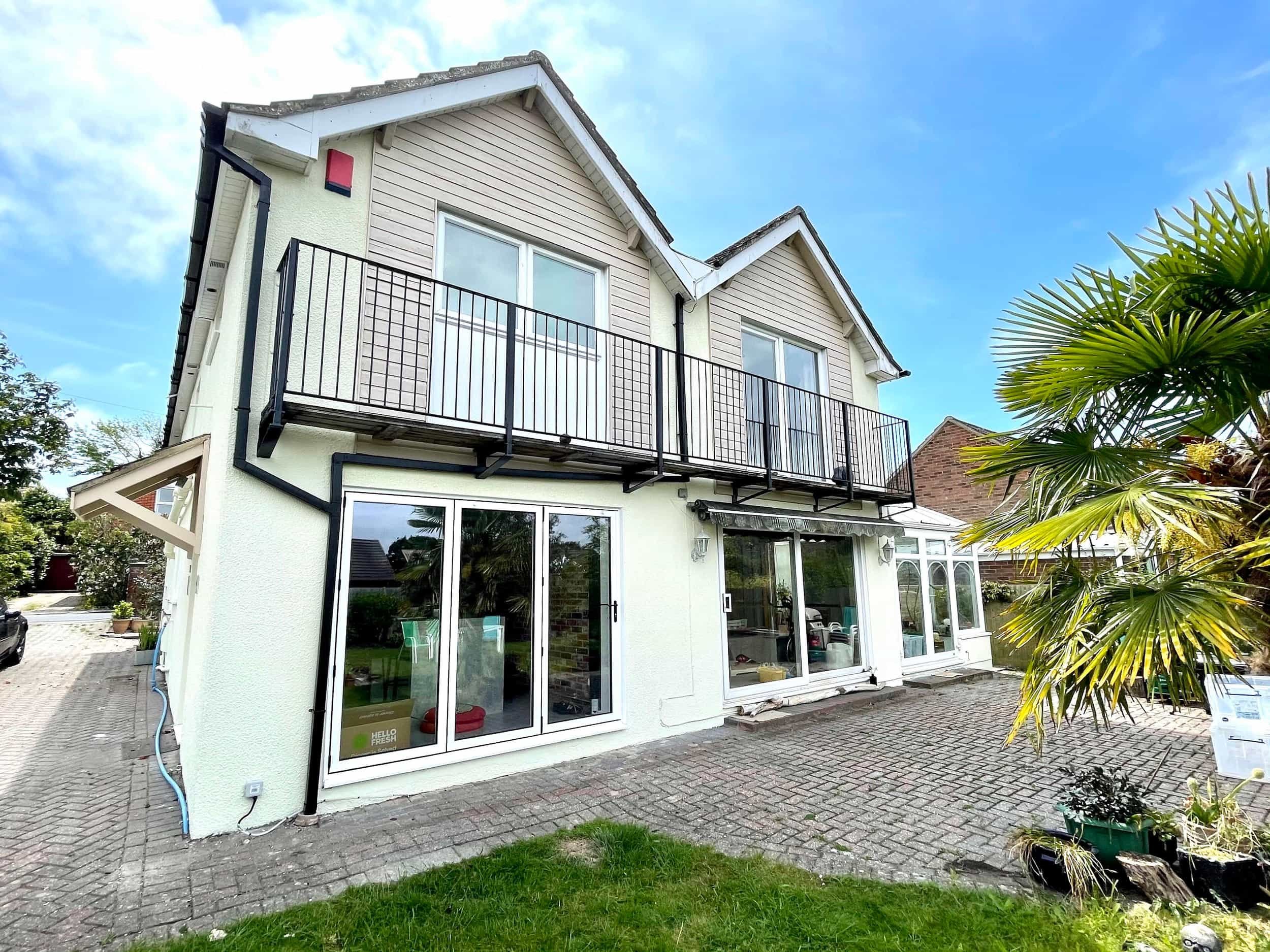 A multi-story house with large sliding glass doors on the ground floor and a balcony on the upper floor. The house has beige siding and a white exterior wall, with a tiled roof. There is greenery around, including a palm tree on the right, and a paved area in front.