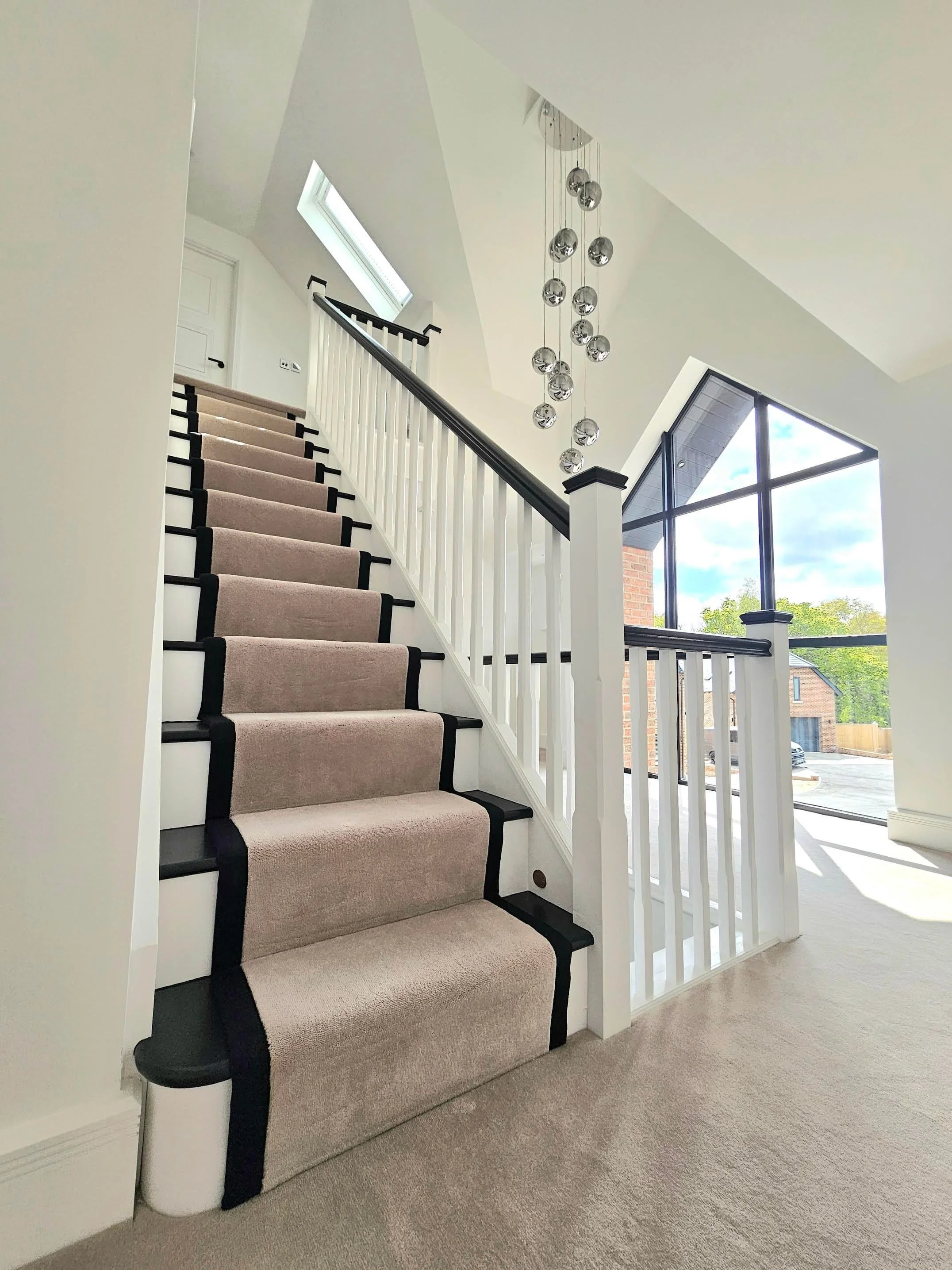 Interior view of a staircase with beige carpet, white railing with black handrail, large windows, and modern chandelier with multiple hanging chrome balls.