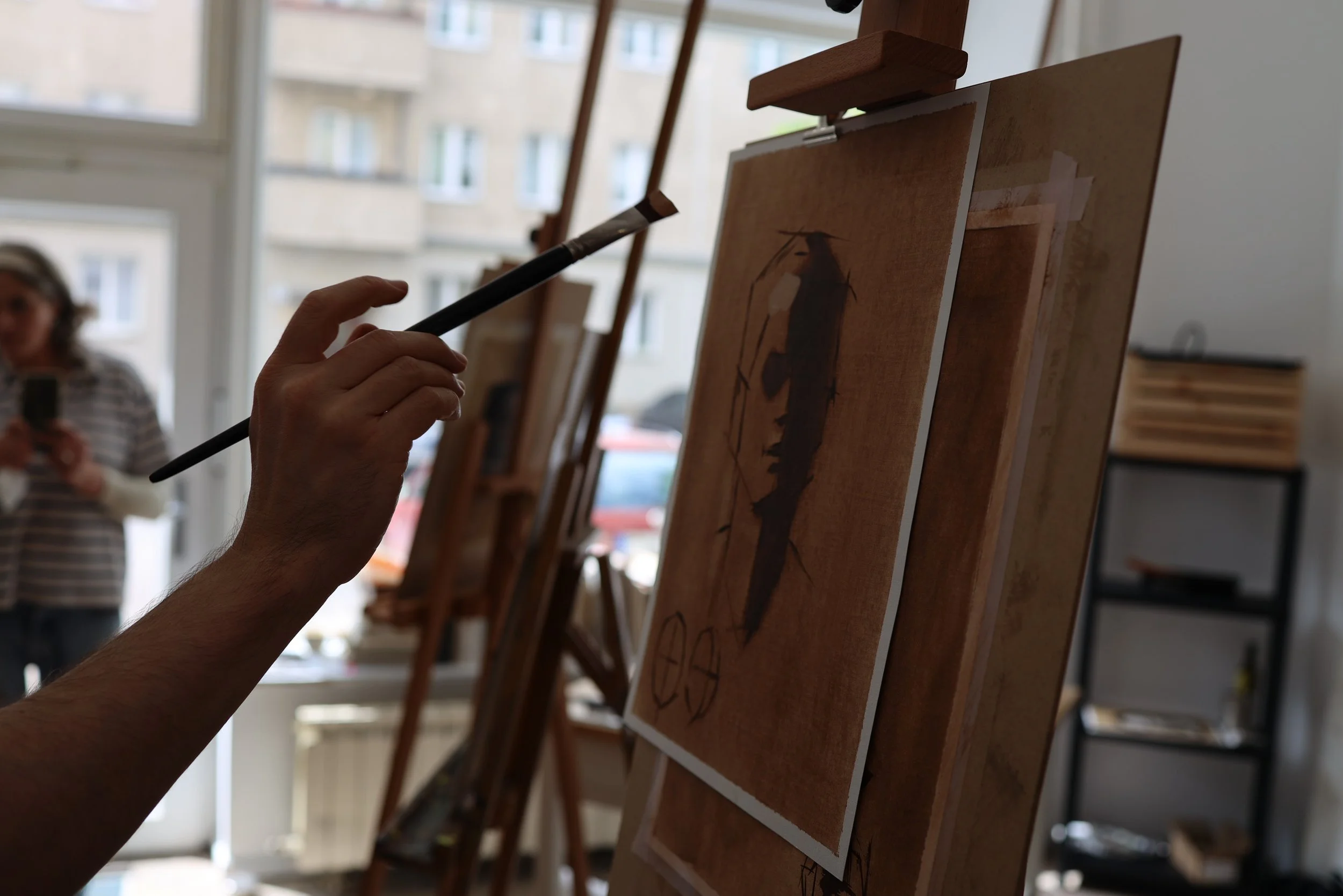 An artist painting a silhouette of a dog on a canvas using a brush in a well-lit art studio with large windows and other canvases on easels.