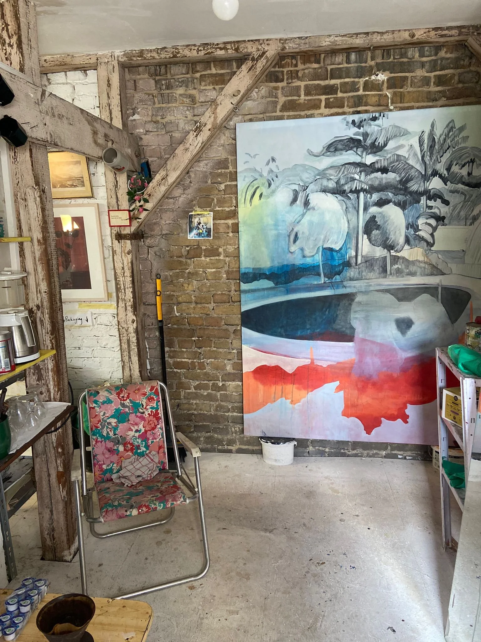 An art studio with a large painting of trees and water, a floral chair, a wooden table with a bowl and cans, a shelf with green bags, and exposed brick walls and wooden beams.