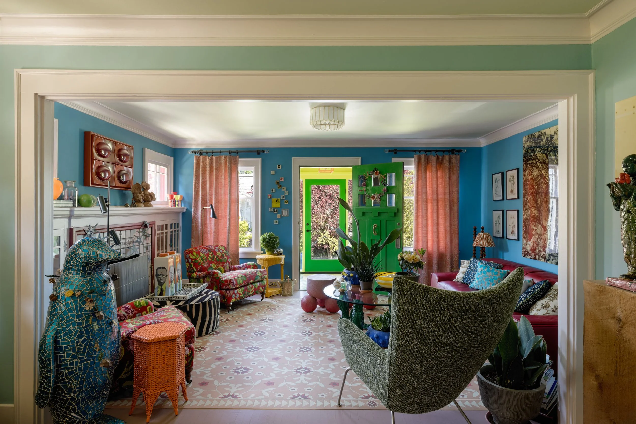 Colorful living room with bright blue walls, pink curtains, and various patterned furniture including a sofa, armchair, and swivel chair. Green door, plants, artwork, and decorative items are visible.