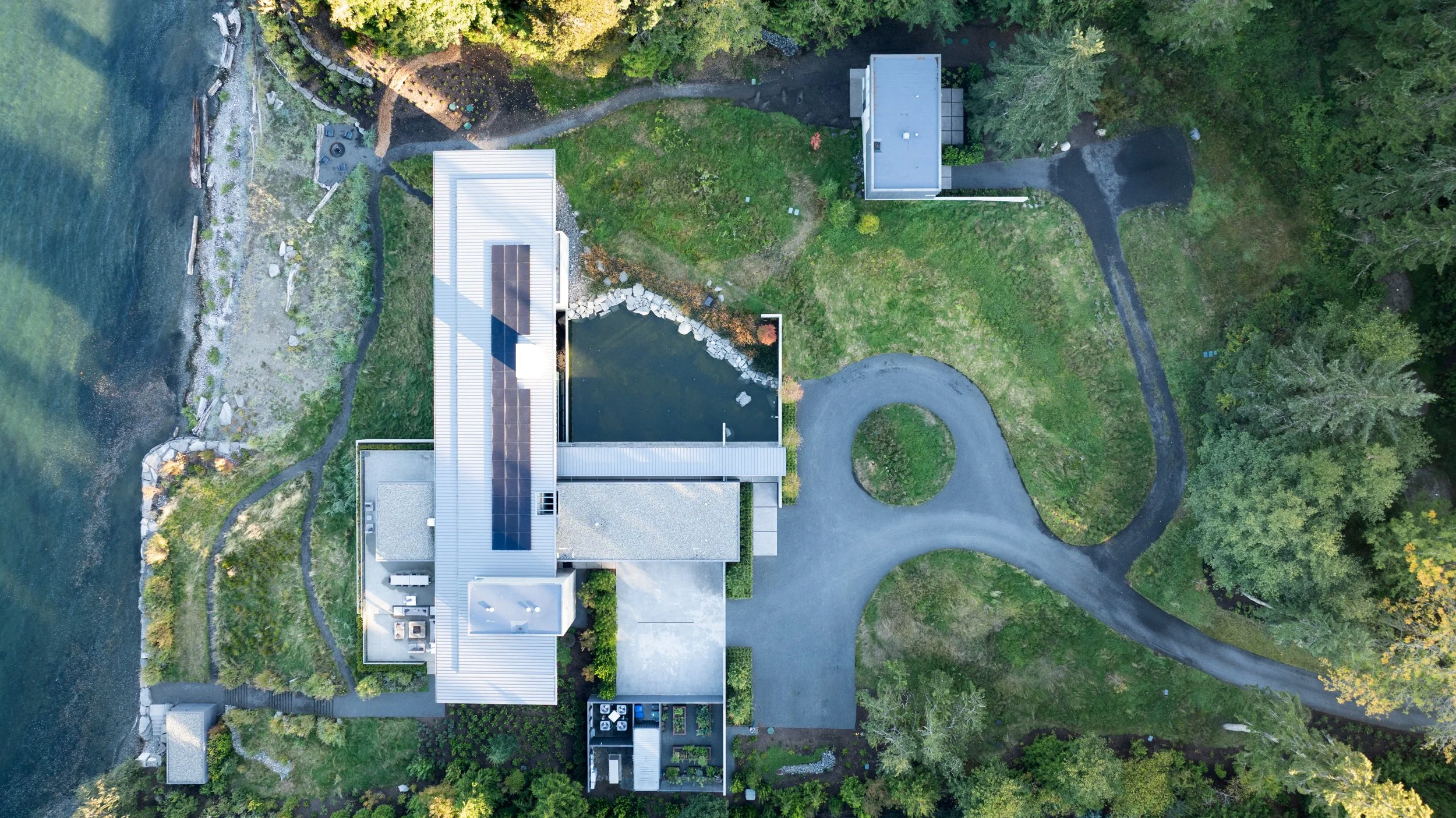 An aerial view of a house with solar panels on the roof, surrounded by greenery, a driveway, a pond, and trees.