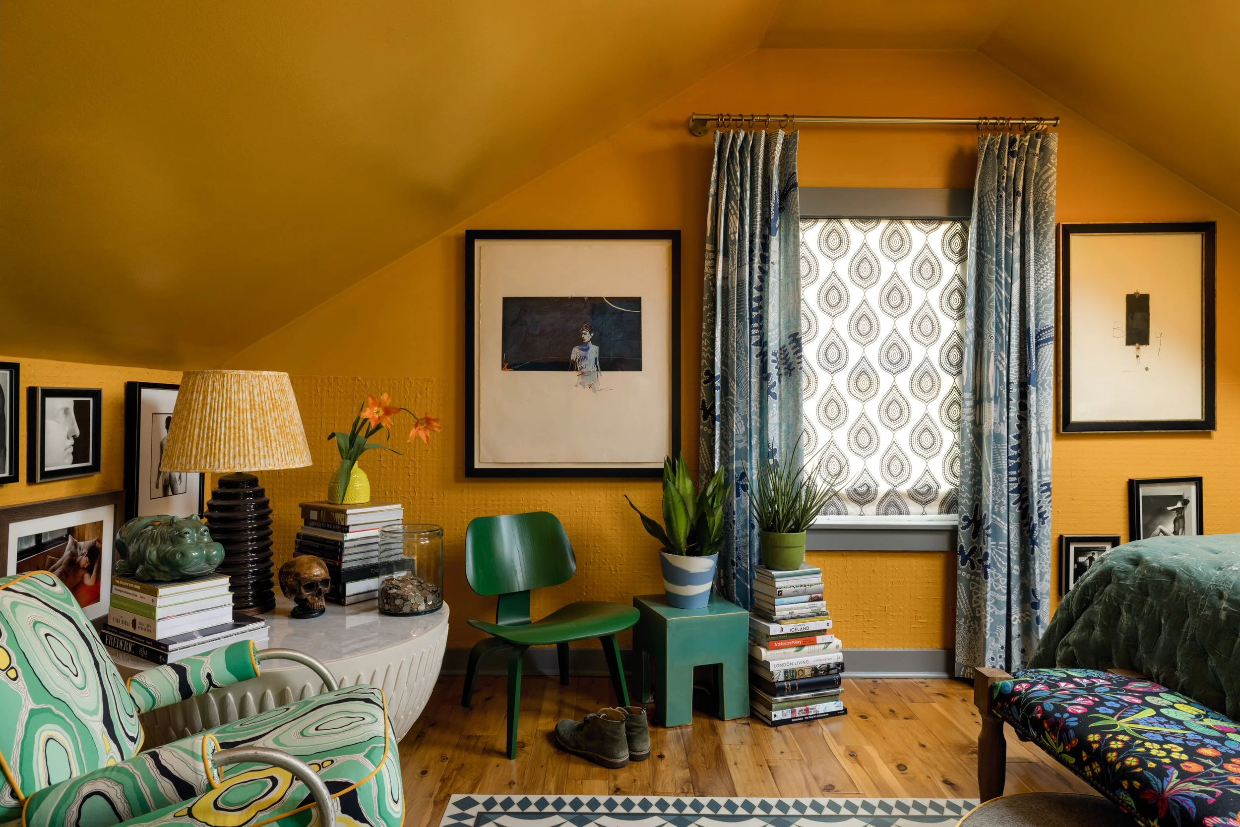 A cozy bedroom with yellow walls and wooden flooring, featuring framed art and photographs on the walls, a window with patterned curtains, a green chair, a small green table with plant pots, stacked books, a bed with a colorful floral blanket, a white marble-top side table with books, a lamp, flowers, and decorative items.