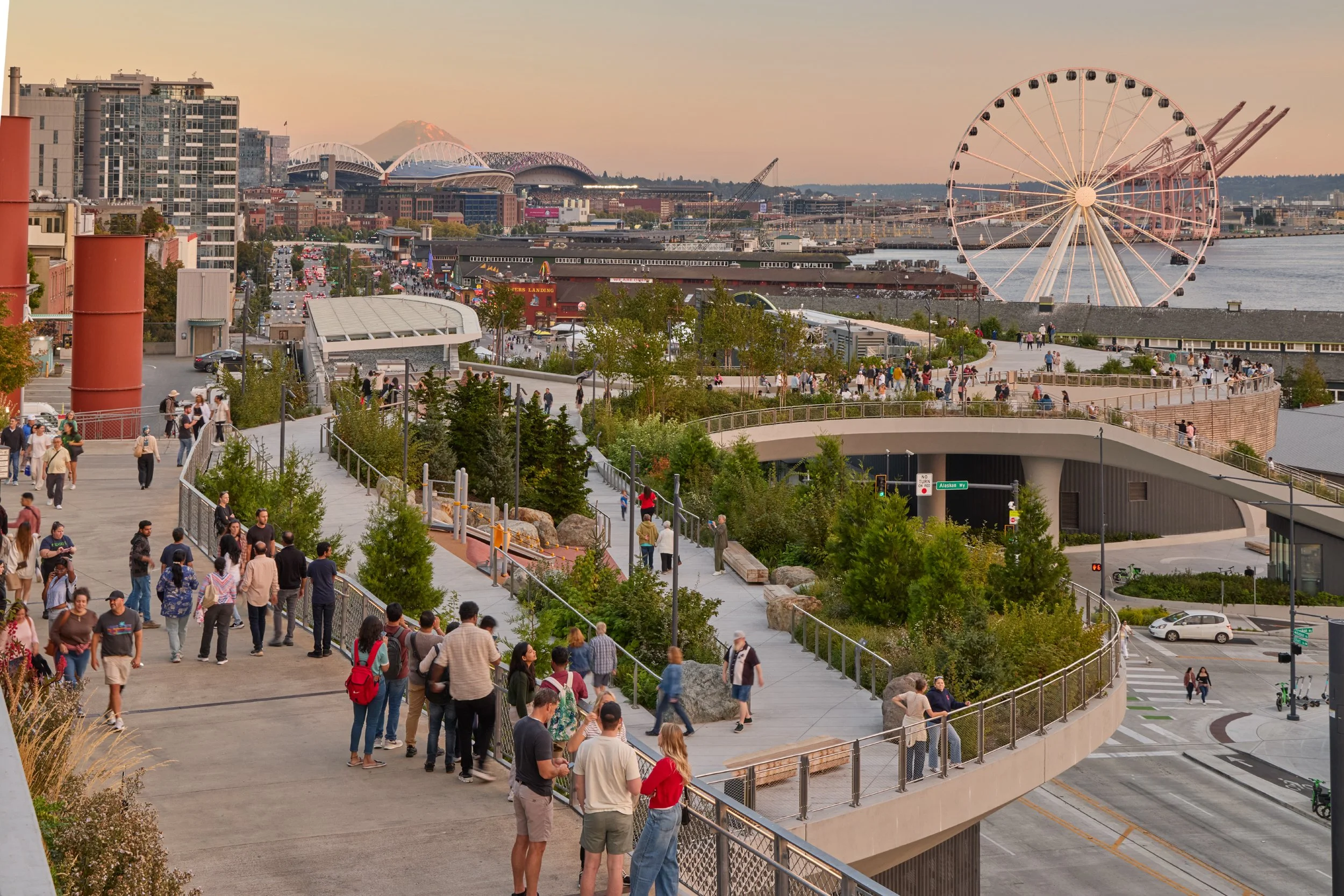 People walking, children playing, and trees on Overlook Walk, a waterfront park with The Great Wheel, boats, and industrial buildings in the background.