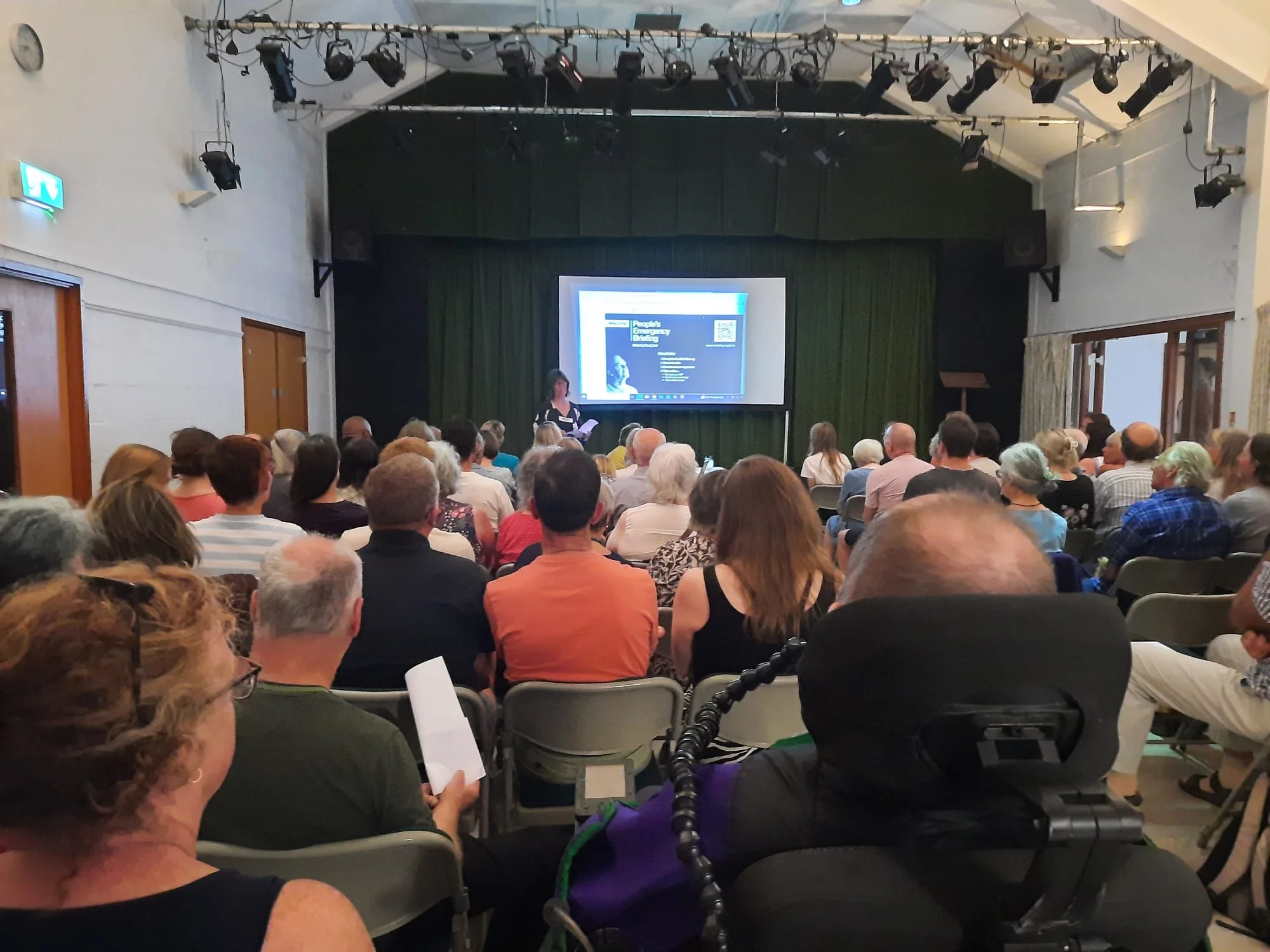 A woman stands in front of a screen addressing a packed audience in a village hall.