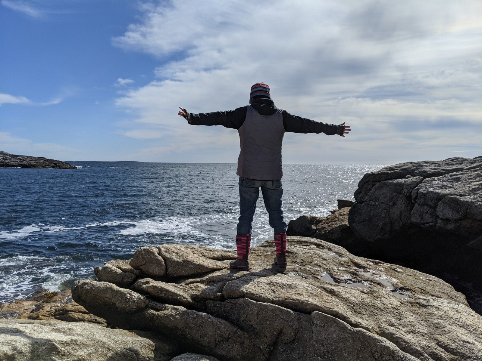 Katherine Grigg standing on the rocky coast of Maine, arms out stretched, facing the ocean.