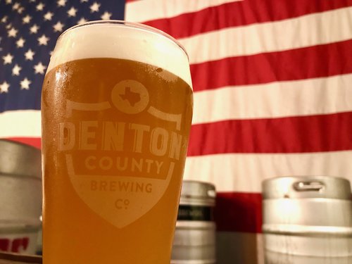 a glass of beer with the denton county brewing company logo sitting on a table in front of beer kegs and the american flag.JPG