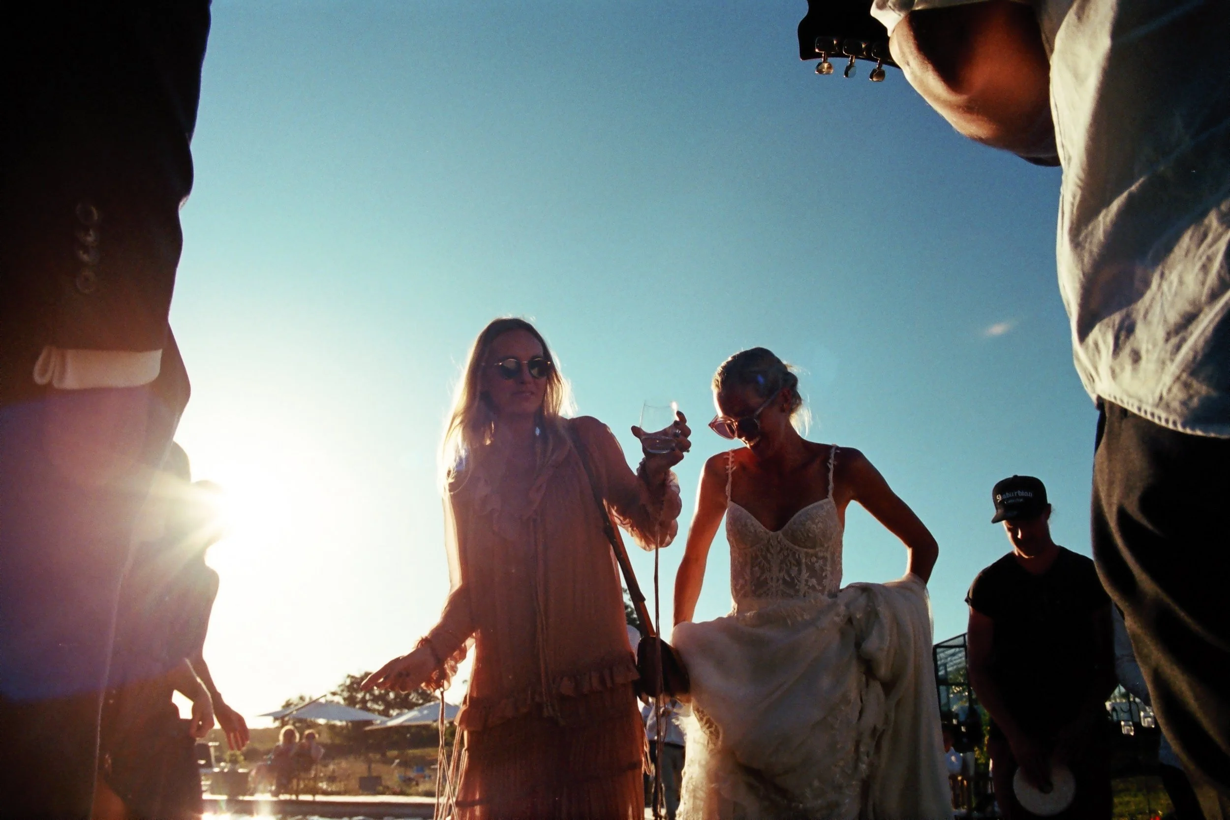 Group of people at an outdoor event during sunset, with two women in the center, one holding a glass of wine and the other in a wedding dress, surrounded by others.