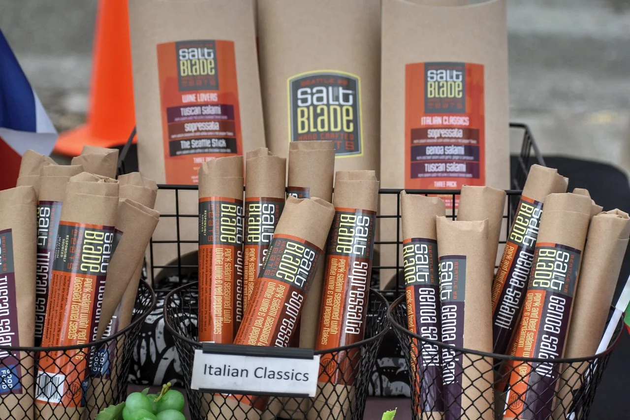 Baskets of Salt Blade handcrafted salami — Tuscan, soppressata, and Genoa — with three-pack gift boxes behind