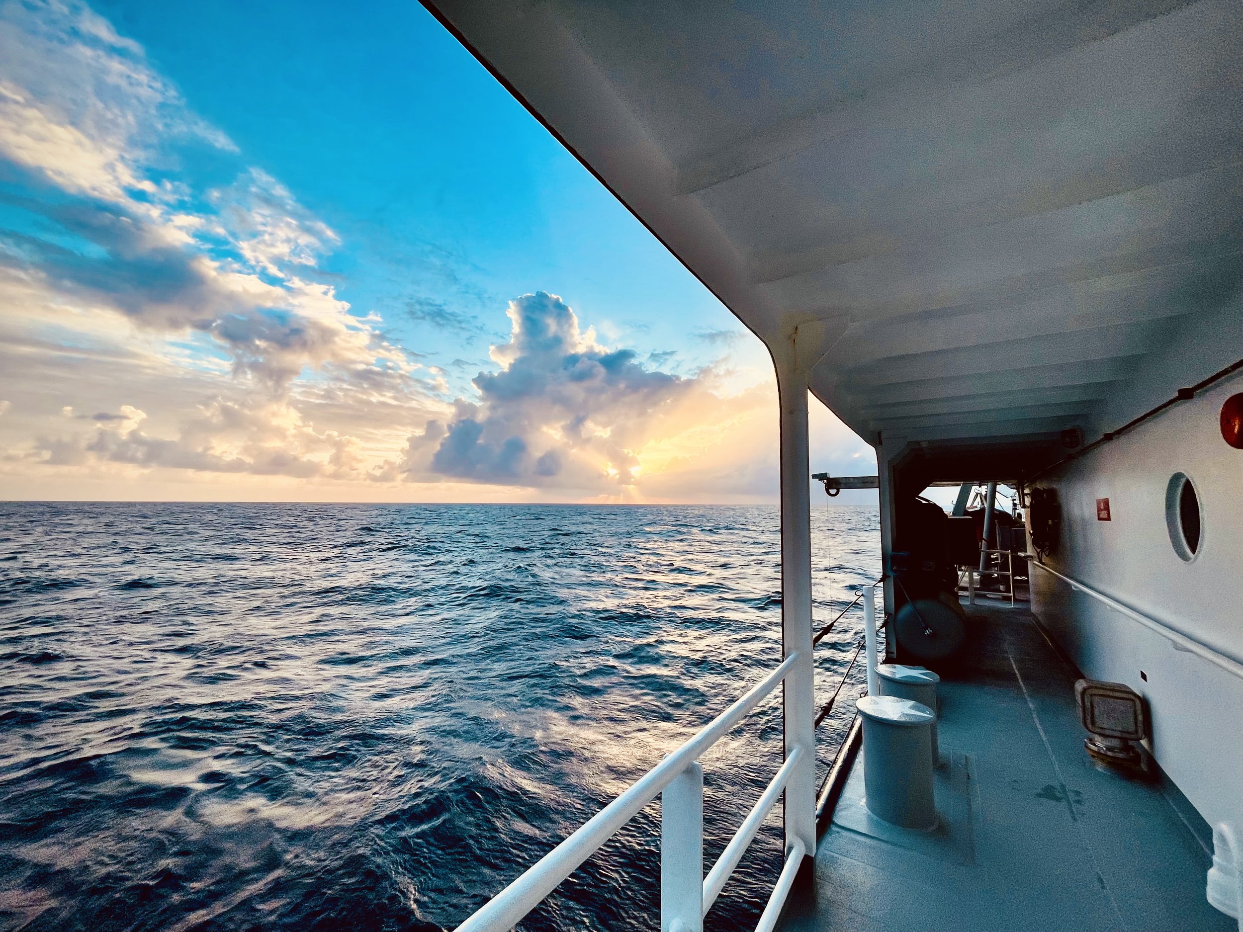 Sunrise over the open Pacific viewed from the side deck of a research vessel, with calm blue water, dramatic clouds, and the ship's railing leaning toward the horizon.