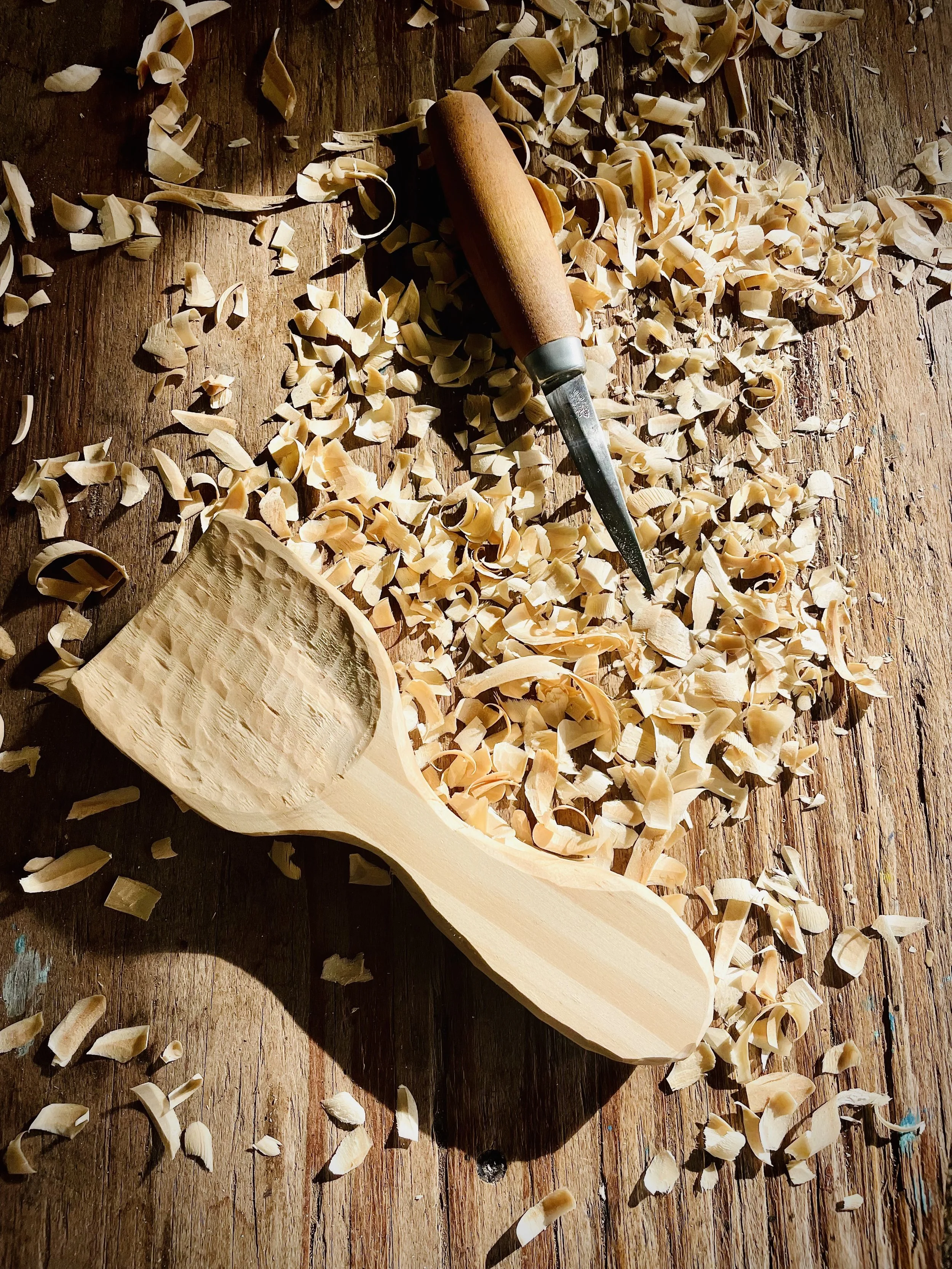 Wooden spoon being carved on a workbench surrounded by wood shavings.