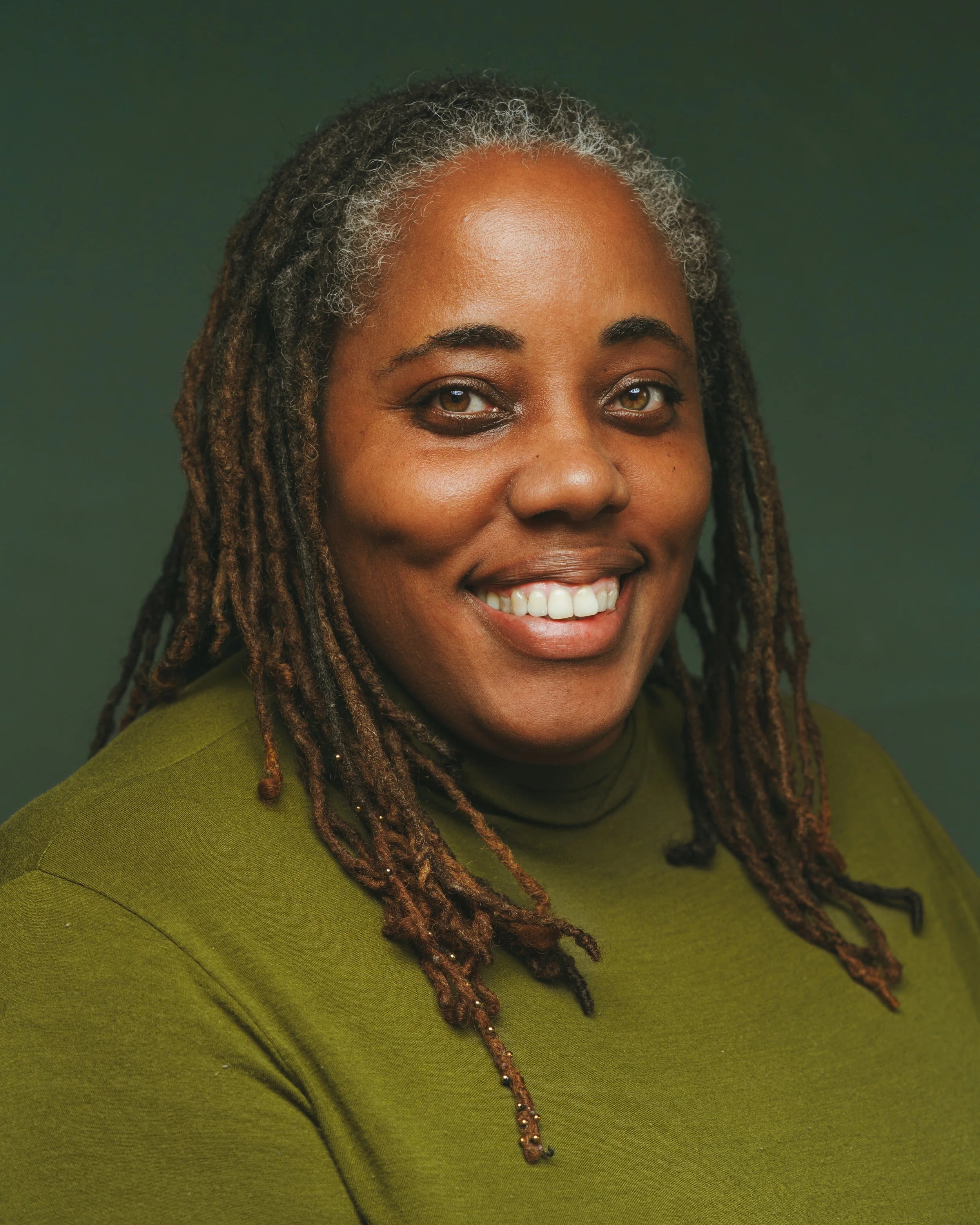 Close-up of a woman with brown skin, gray and brown dreadlocks, smiling, wearing a green top, against a green background.