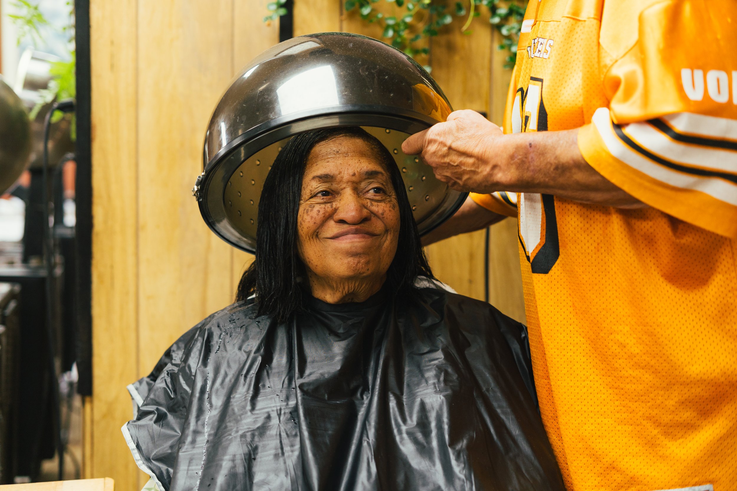A woman with dark hair getting her hair washed at a salon, with a black hair dryer hood over her head, and a barber in a yellow sports jersey working on her hair.