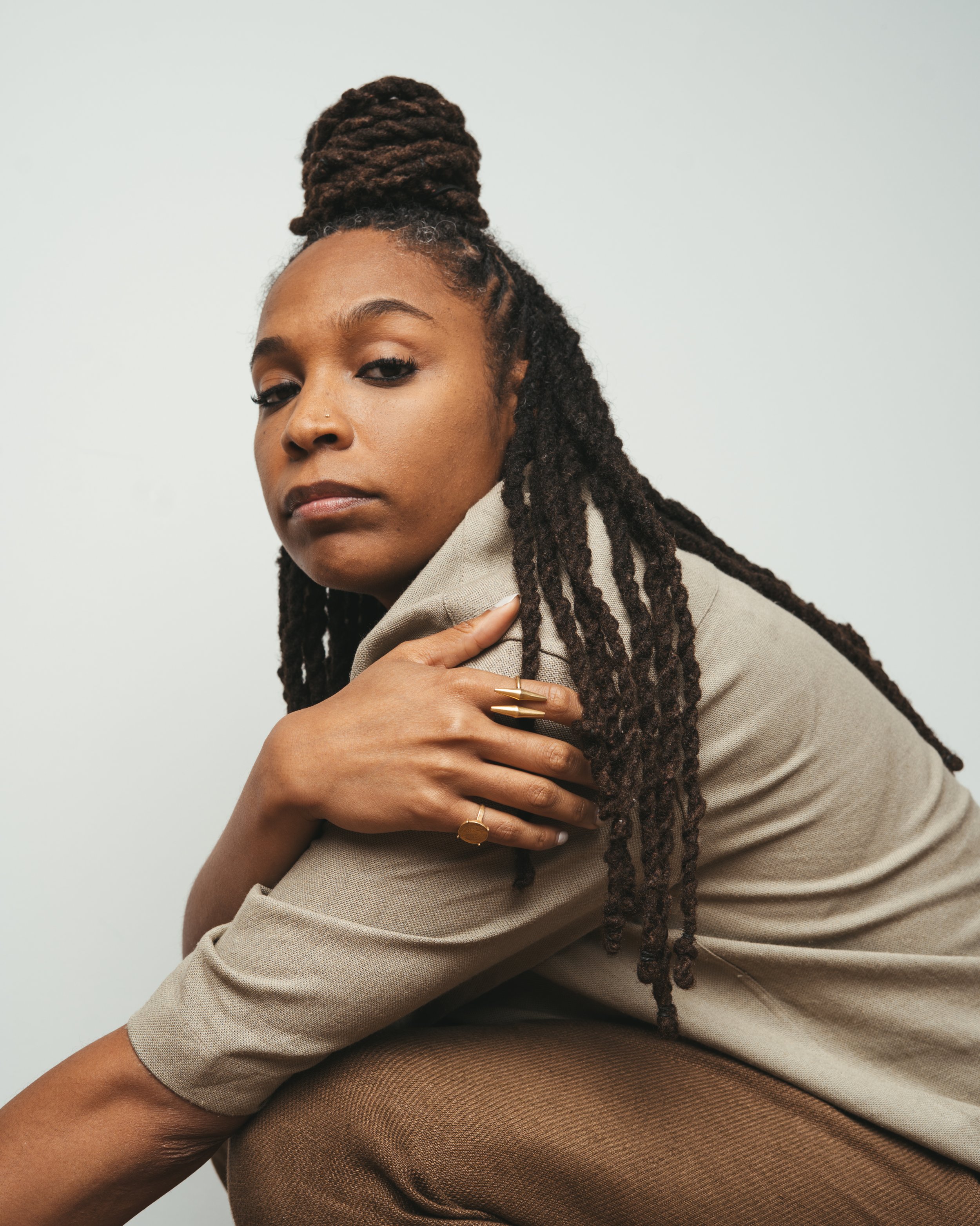 Close-up of a woman with dreadlocks, wearing rings, and a beige blazer, sitting against a plain white background.