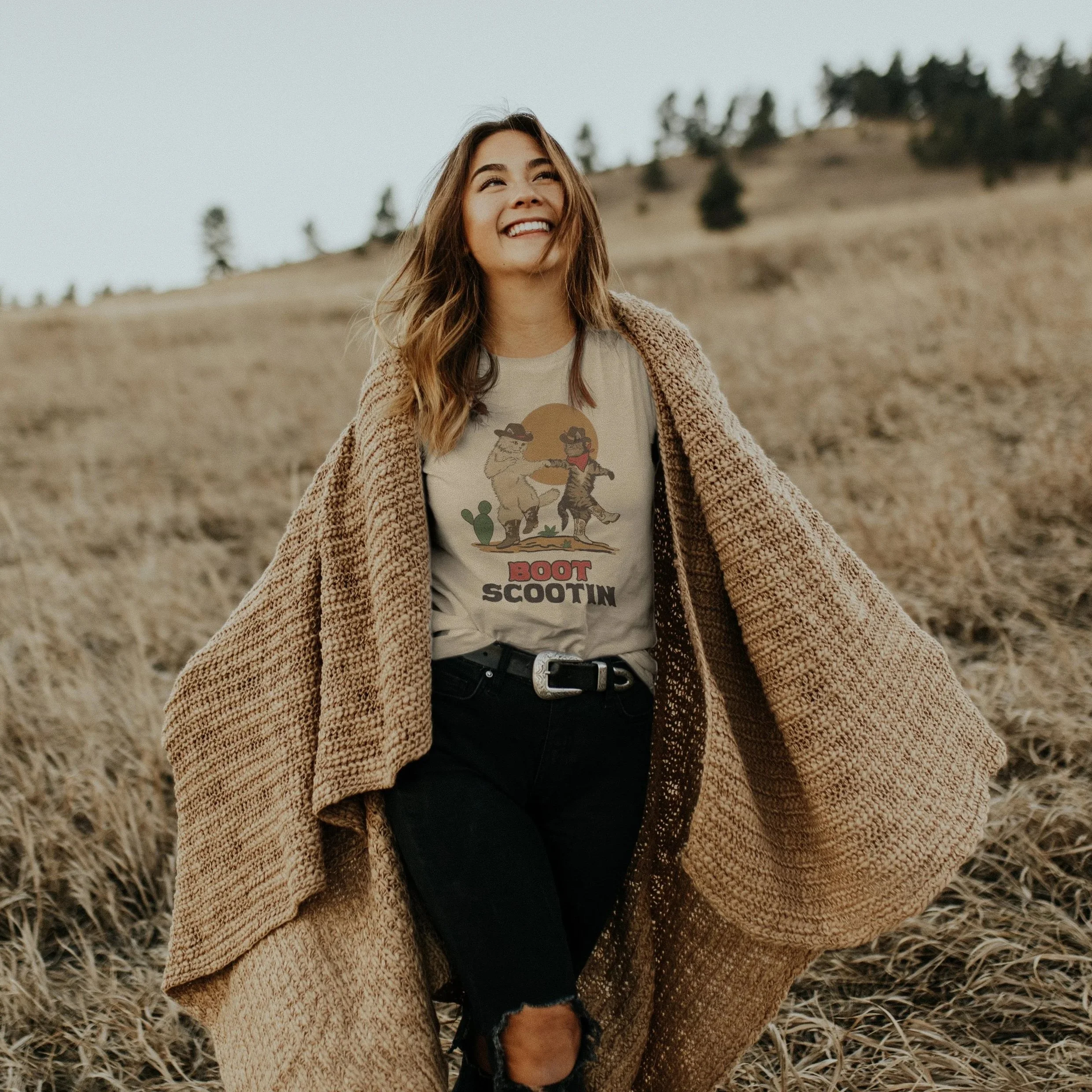 Young woman smiling and walking outdoors in a field, wearing a t-shirt with cartoon characters, a belt, ripped jeans, and a large brown coat.