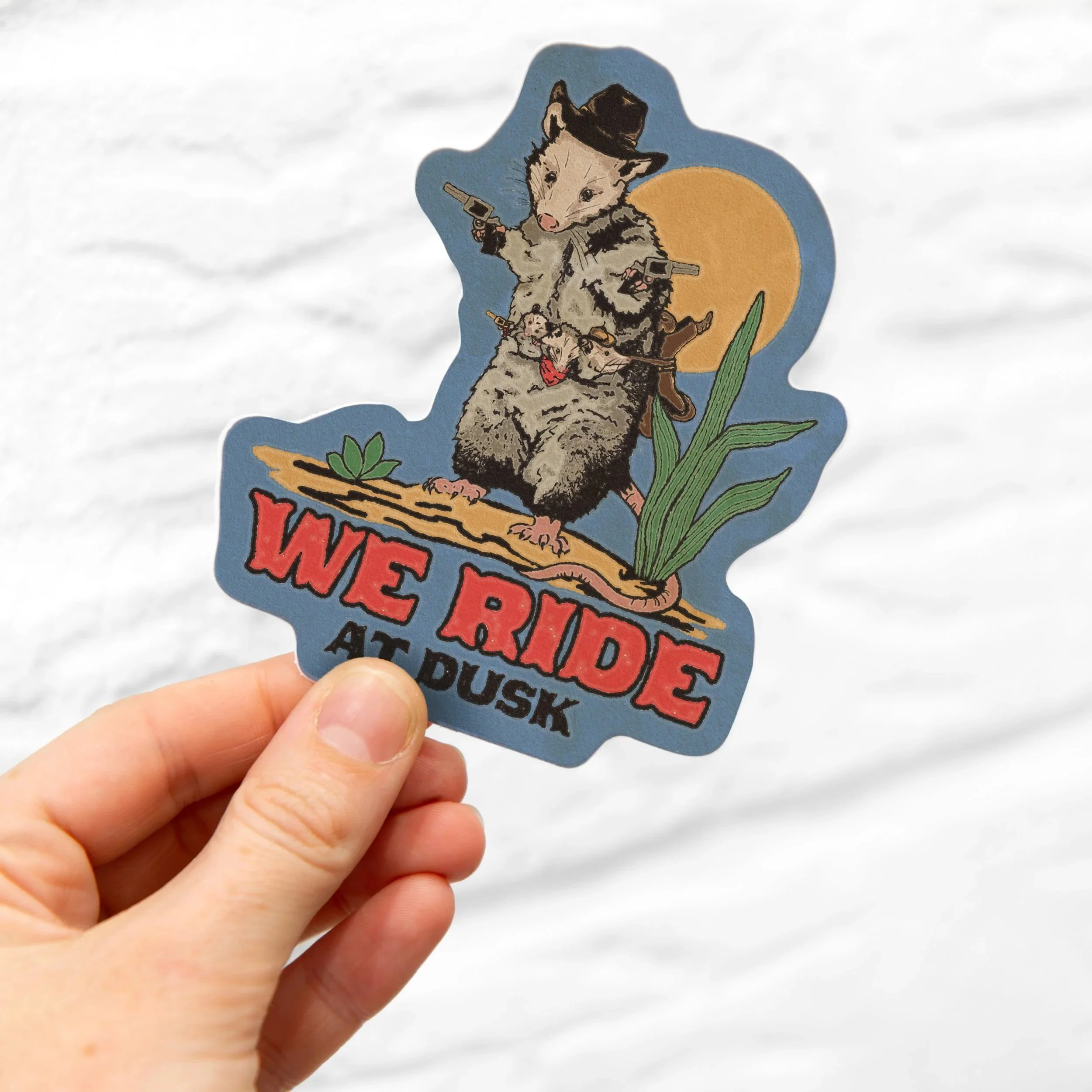 A sticker showing a raccoon dressed as a cowboy, holding guns, riding a snake, with the text 'WE RIDE AT DUSK'.