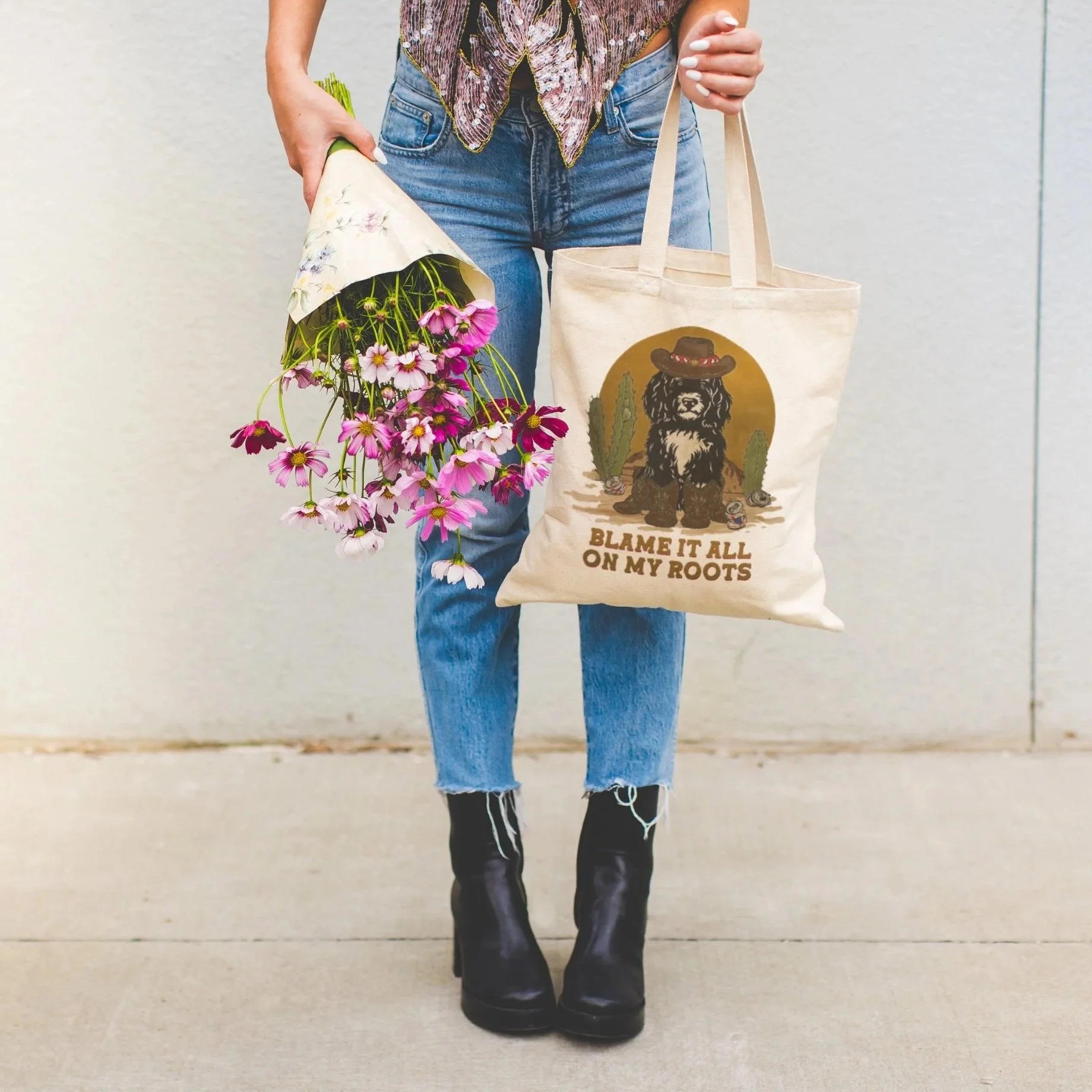 Person holding a bouquet of pink and purple flowers, carrying a tote bag with a bear wearing a hat, cactus, and the text "Blame it all on my roots". The person is wearing blue jeans and black boots.