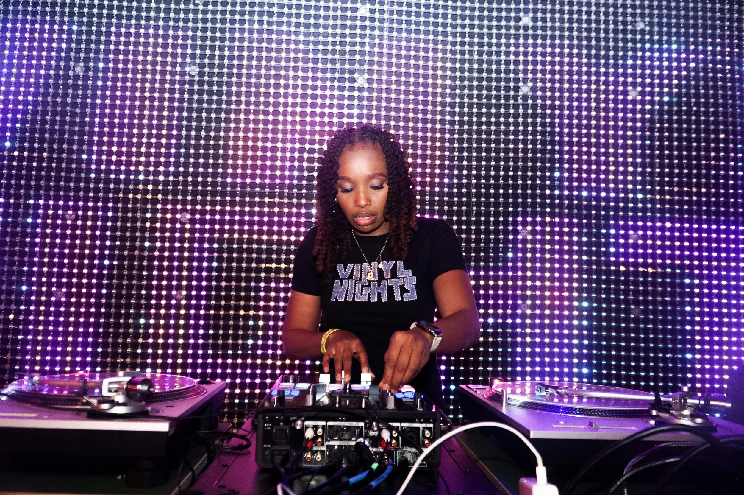 A woman DJing with turntables and a mixer in front of a colorful LED backdrop, wearing a black shirt with 'VYNL NIGHTS' printed on it, and looking down at her equipment.
