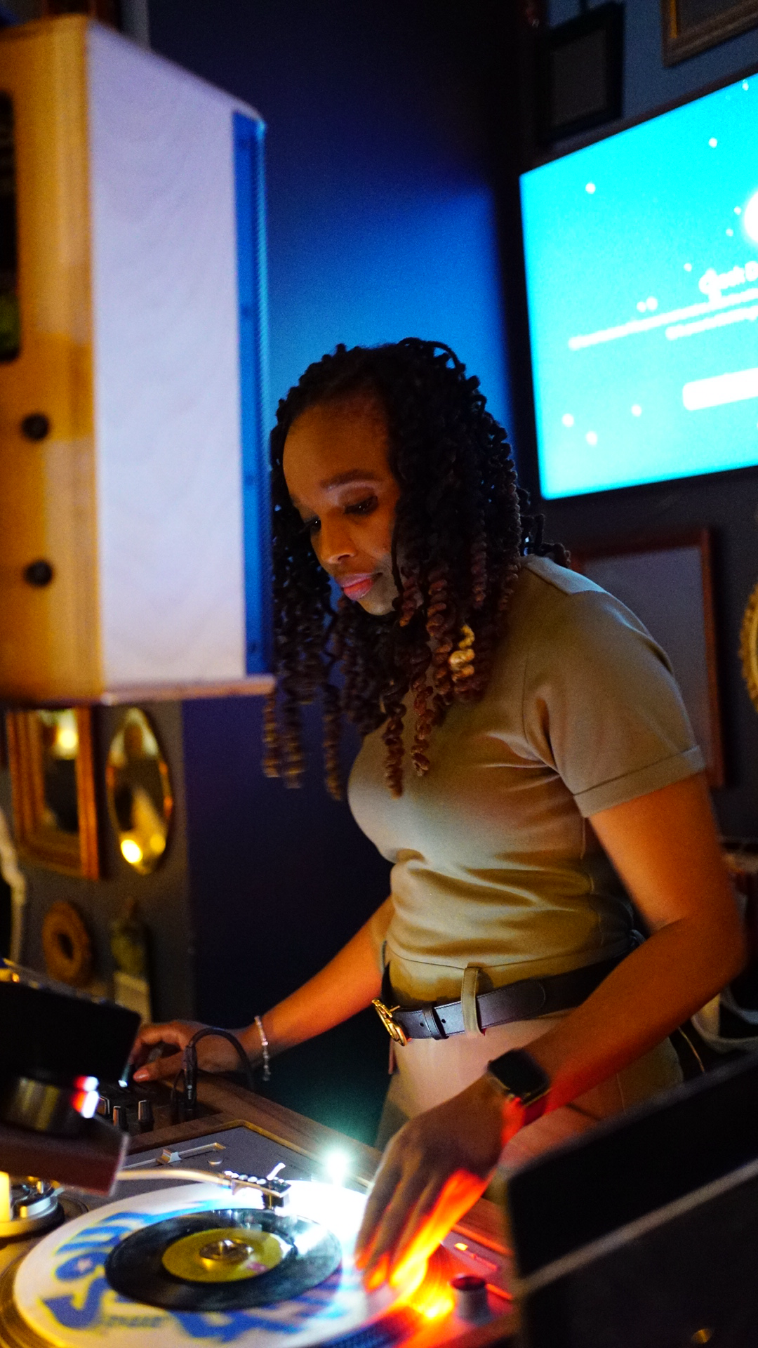 A woman with curly hair DJing at a club or party, with a blue screen in the background.