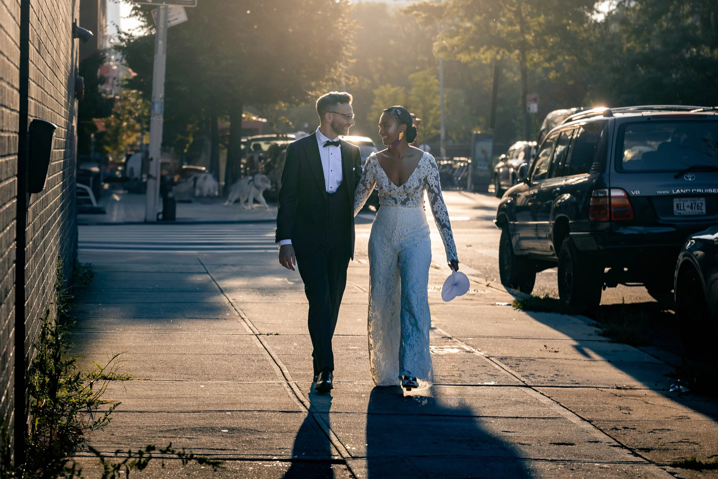A couple dressed in wedding attire walking on a city sidewalk, smiling at each other, with sunlight shining through trees and parked cars along the street in the background.