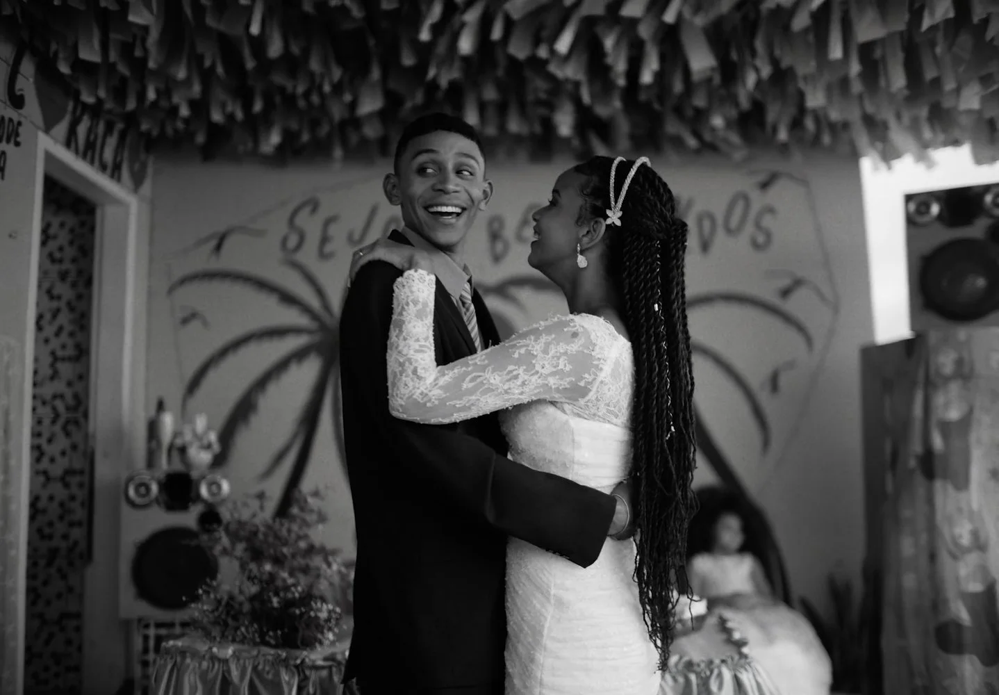 A smiling couple, the man in a suit and the woman in a wedding dress, dance together at a wedding reception with decorations and a mural in the background.