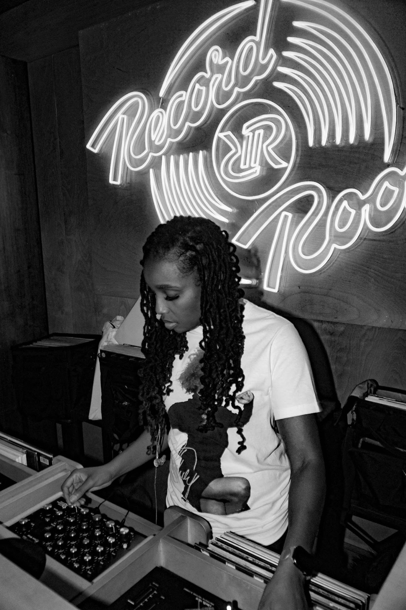 A woman with long, braided hair is standing at a DJ booth, looking down and operating equipment. Behind her is a neon sign that reads 'Radio' with a logo involving the letters R and D. The setting appears to be a record store or a club with shelves o