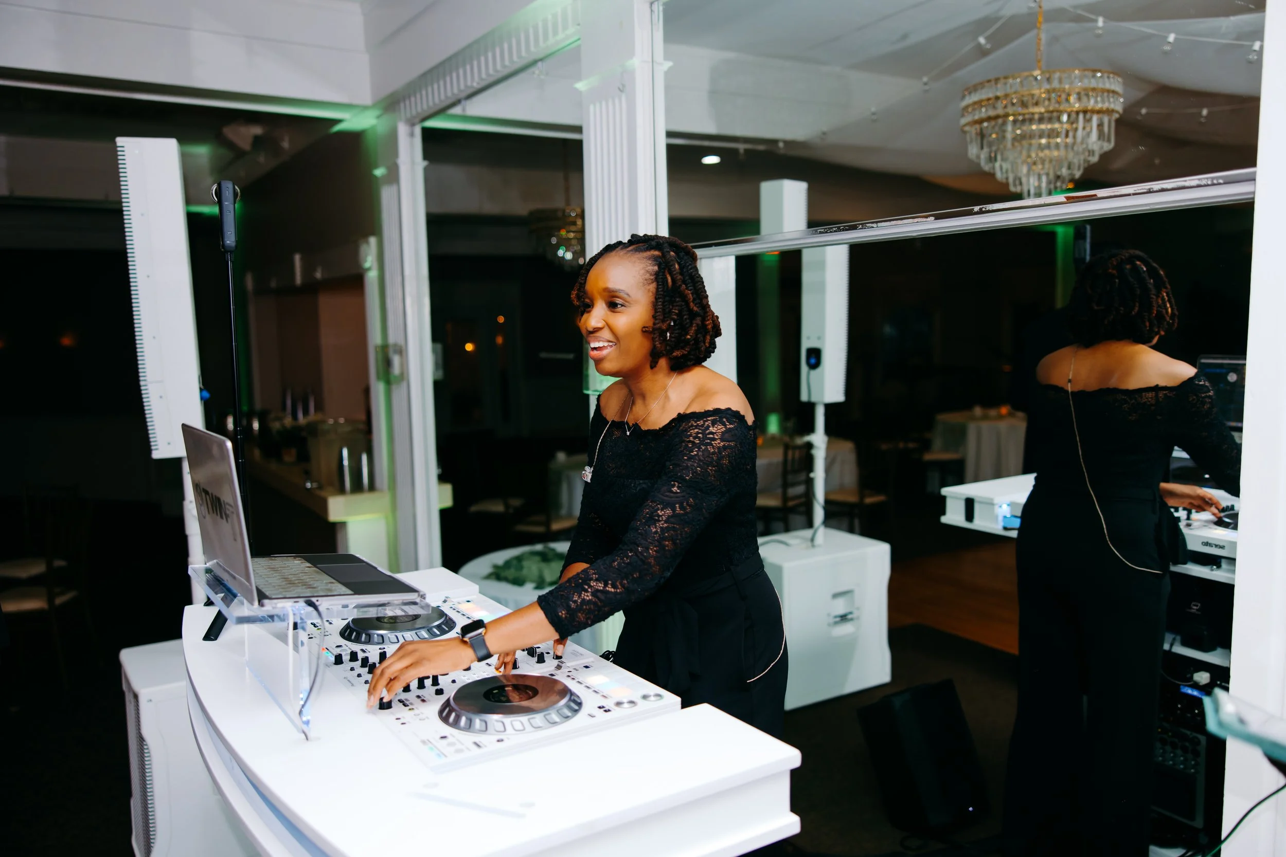 A woman with short curly hair, wearing a black lace top is DJing at an event, standing behind a white DJ console with equipment. In the background, there is another person operating DJ equipment and a chandelier hanging from the ceiling.