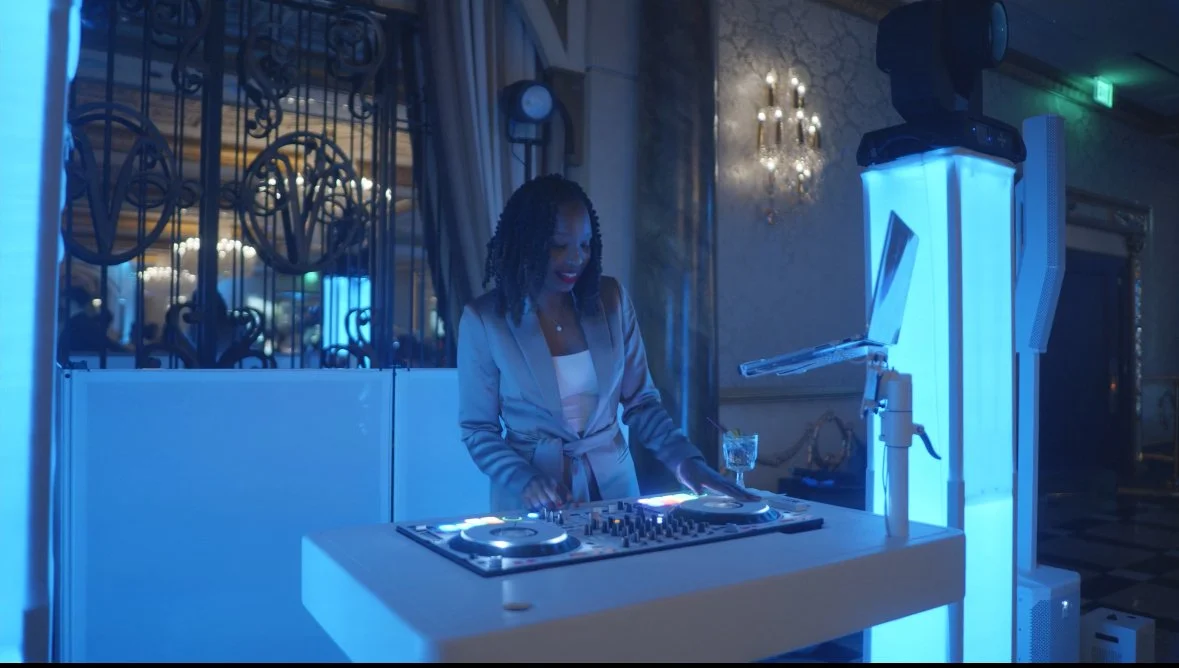 Female DJ in a formal gray suit with curly hair mixing music at a dance event, illuminated by blue lights, in a decorated event space with chandeliers and ornate wall decor.