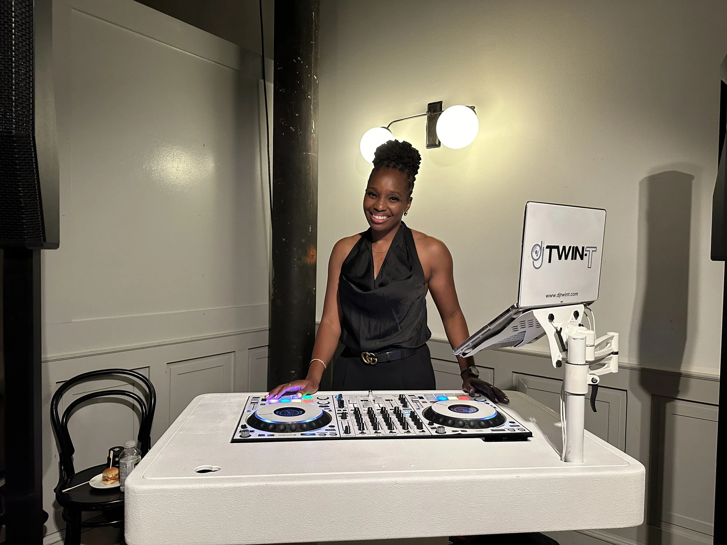 A woman standing behind DJ equipment, smiling, in a room with white walls and a lamp overhead.