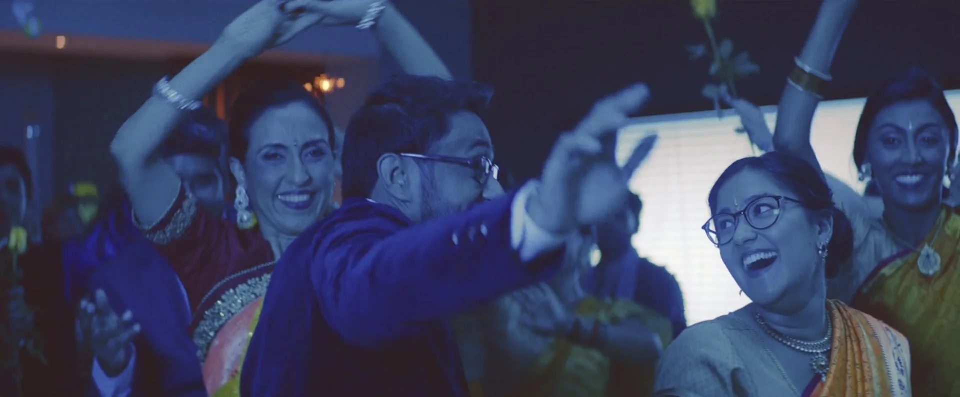 A group of people dancing and celebrating indoors, dressed in traditional Indian clothing, with joyful expressions.