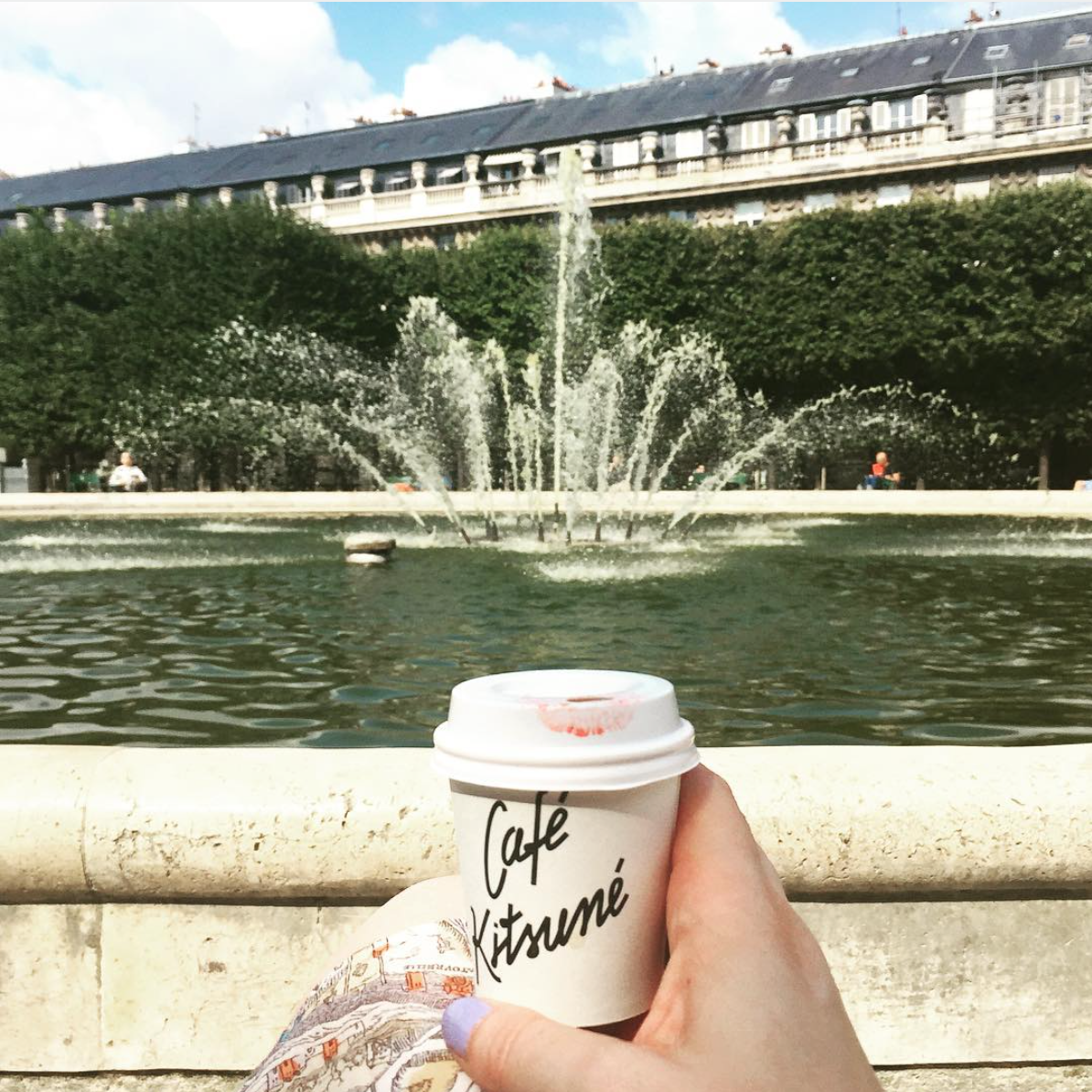 Someone holding an espresso in a paper cup with lipstick on it that says Kitsuné in front of the fountain of the Palais Royal on a sunny day in Paris