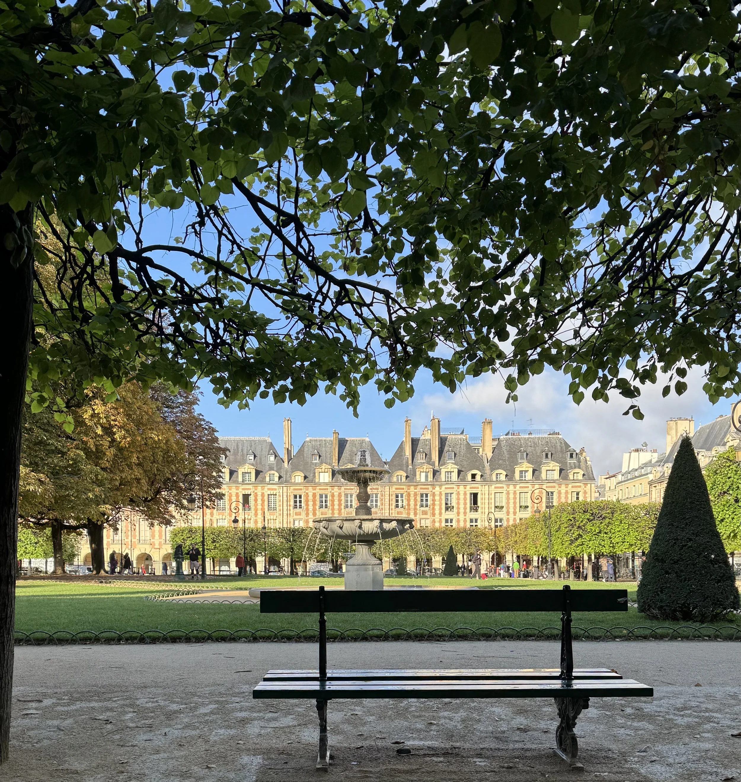 Tree branches fram ethe top of a view on the Places des Vosges with a bench in the foreground with a fountain and the red brick buildings across the square