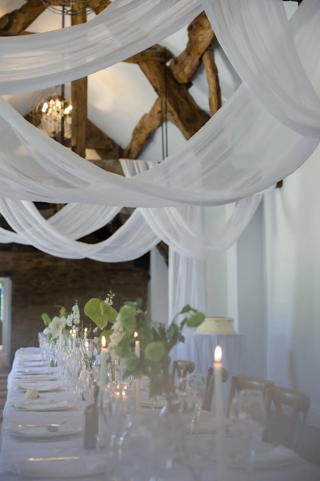 Elegant wedding reception table with white tablecloth, floral arrangements, candles, and a rustic chandelier hanging from wooden beams.