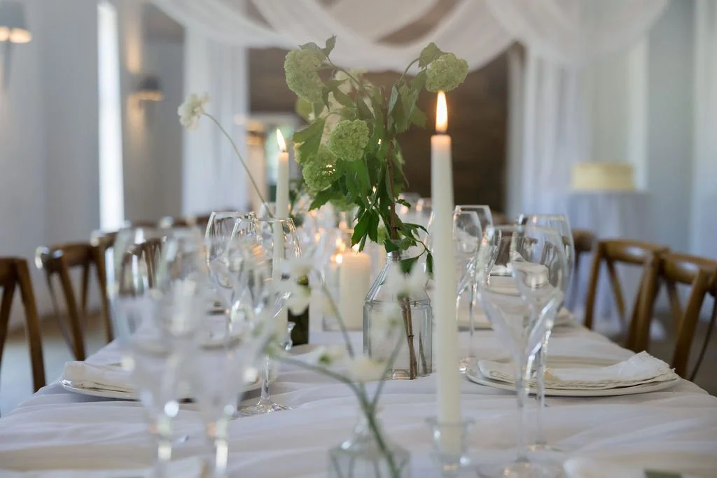 Elegant dining table set with white tablecloth, glassware, white napkins, and tall white candles in a softly lit room with white curtains.
