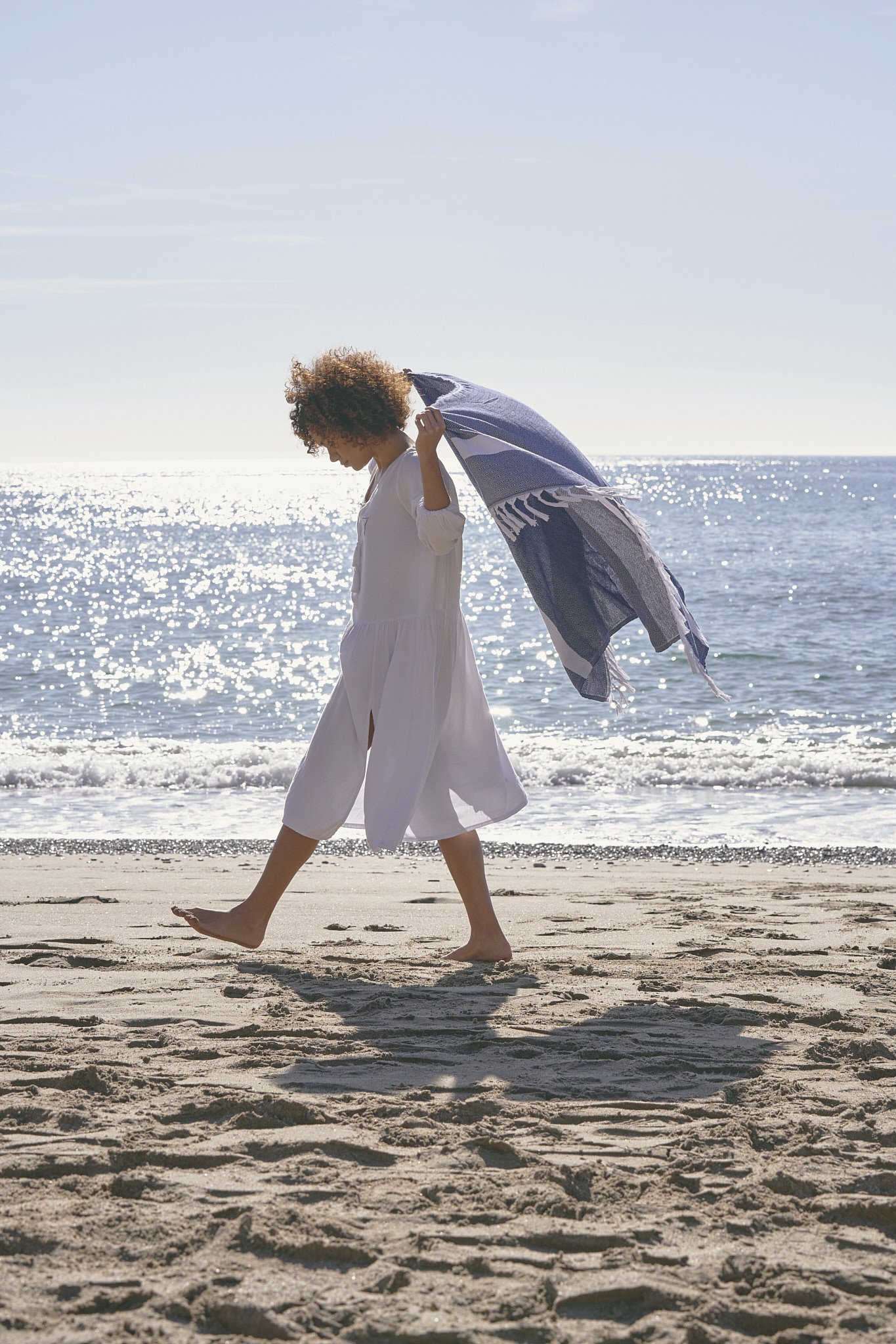 Person walking on beach holding a blue and white towel, wearing a white dress, with the ocean in the background.