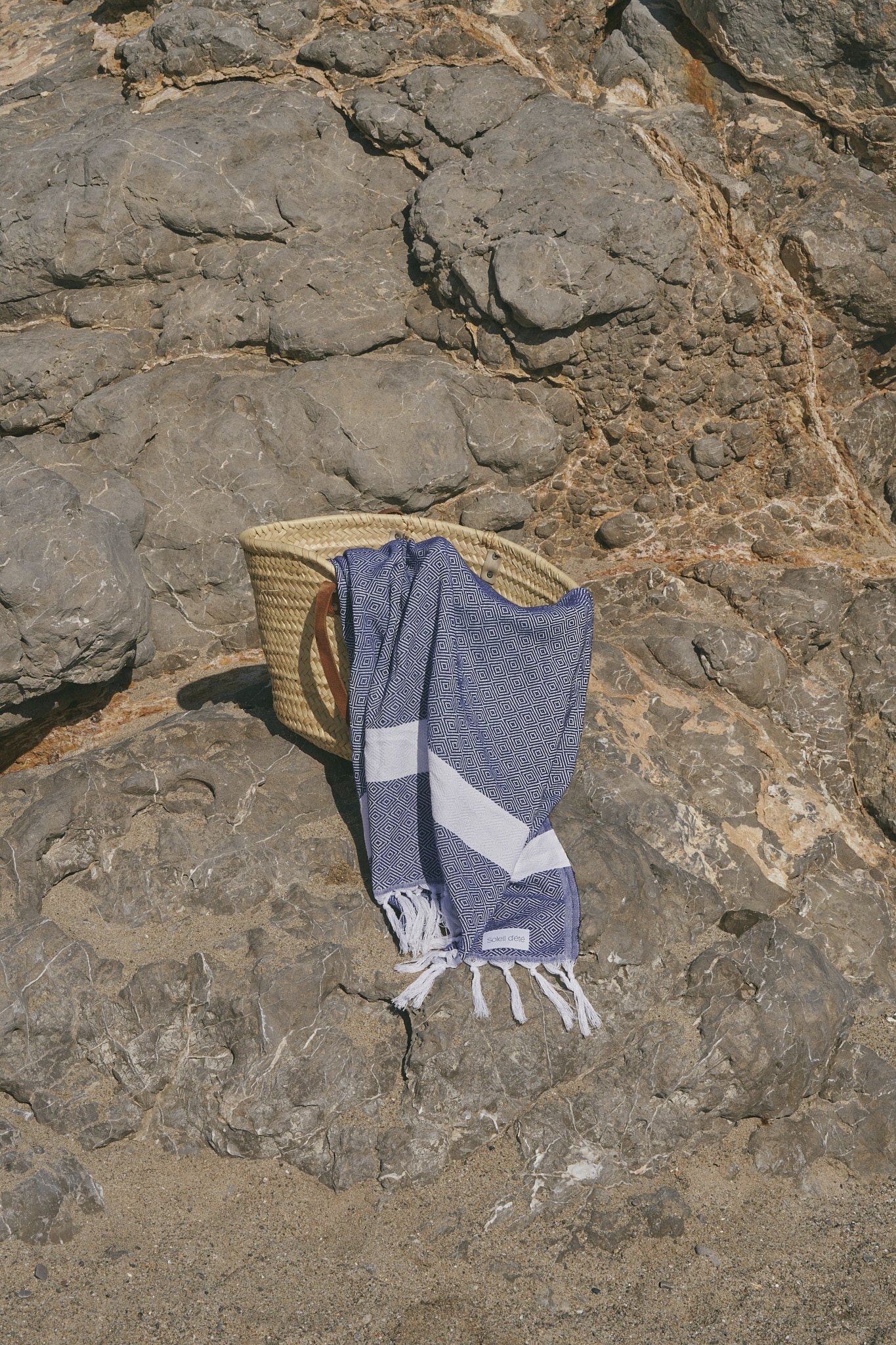 Woven basket with a blue patterned beach towel draped over it, placed on large rocks shot by Barcelona Product and Brand Photographer Julia Malinowska.
