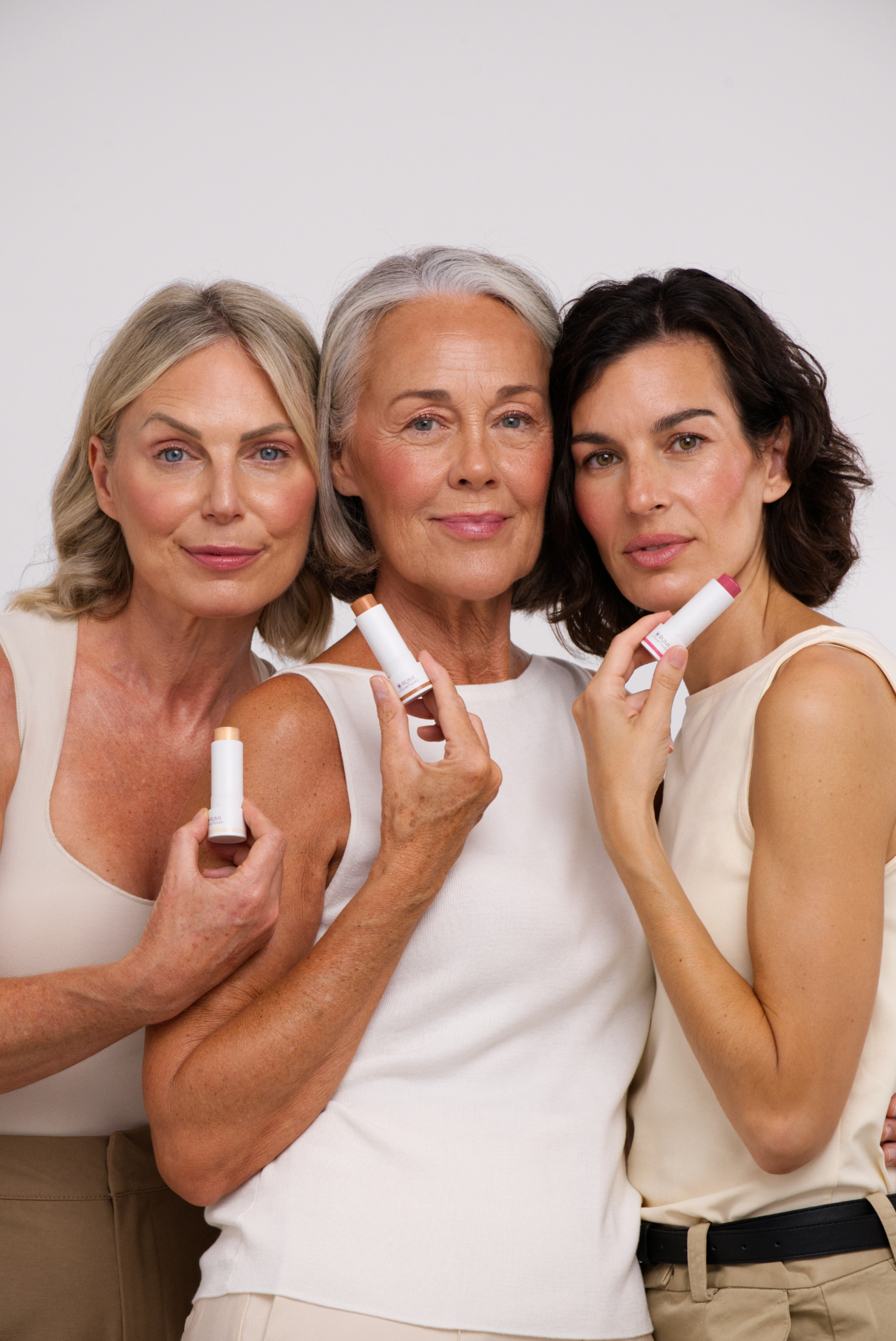 Three women over 40 with glowing, radiant skin showcasing Rumi Cosmetiques beauty sticks in a professional studio beauty photoshoot